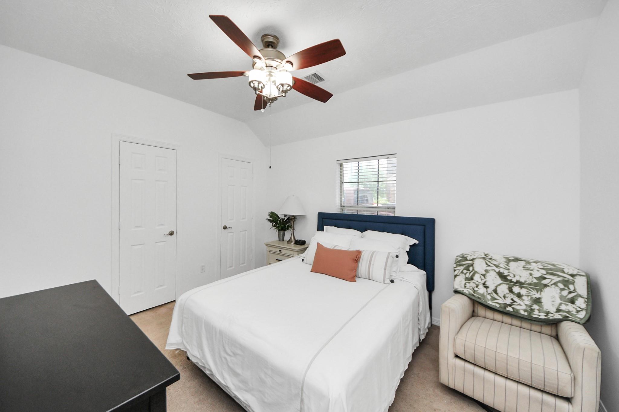 Bedroom with a white bed, navy headboard, ceiling fan, and armchair with a leafy throw.