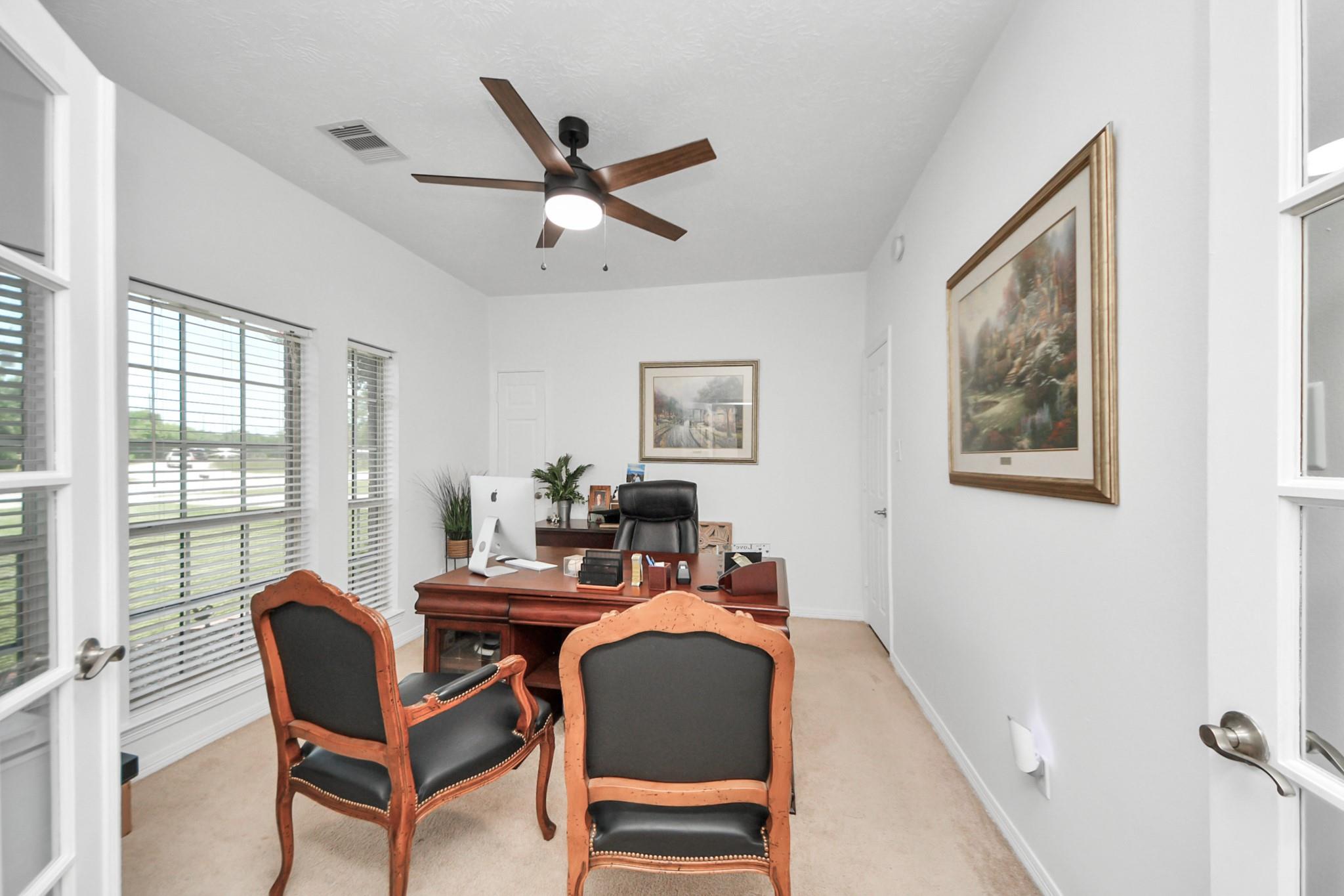 Furnished home office with a wooden desk, two guest chairs, and large windows with blinds.