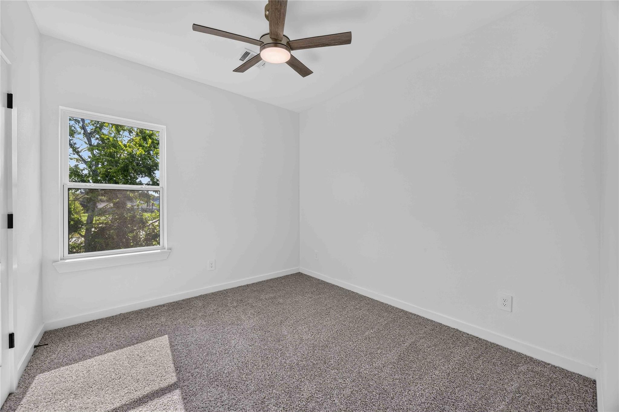 Empty carpeted room with a window overlooking trees and a ceiling fan.