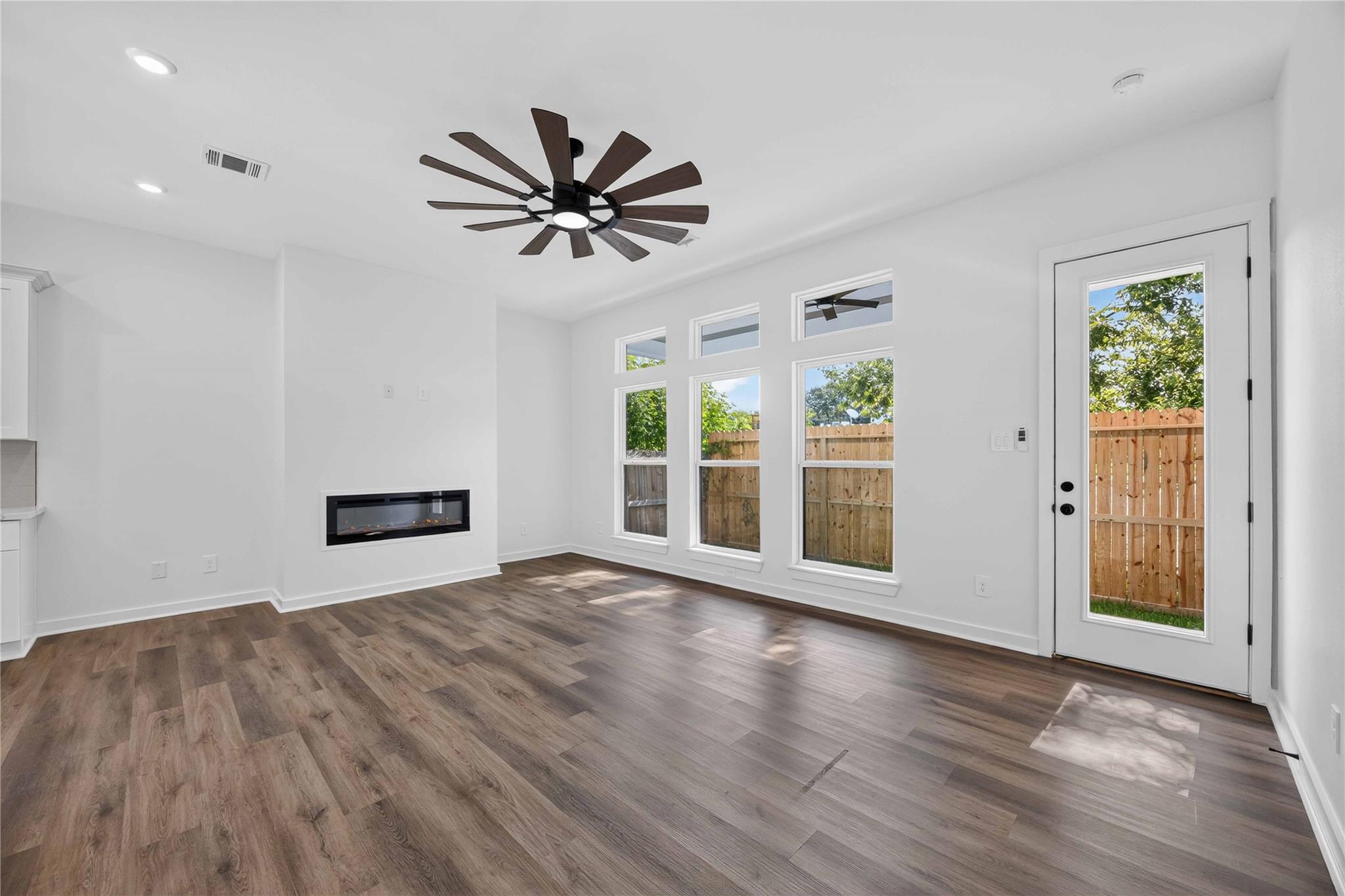 Vacant living room with wood-look floors, linear fireplace, and tall windows overlooking a fenced yard.
