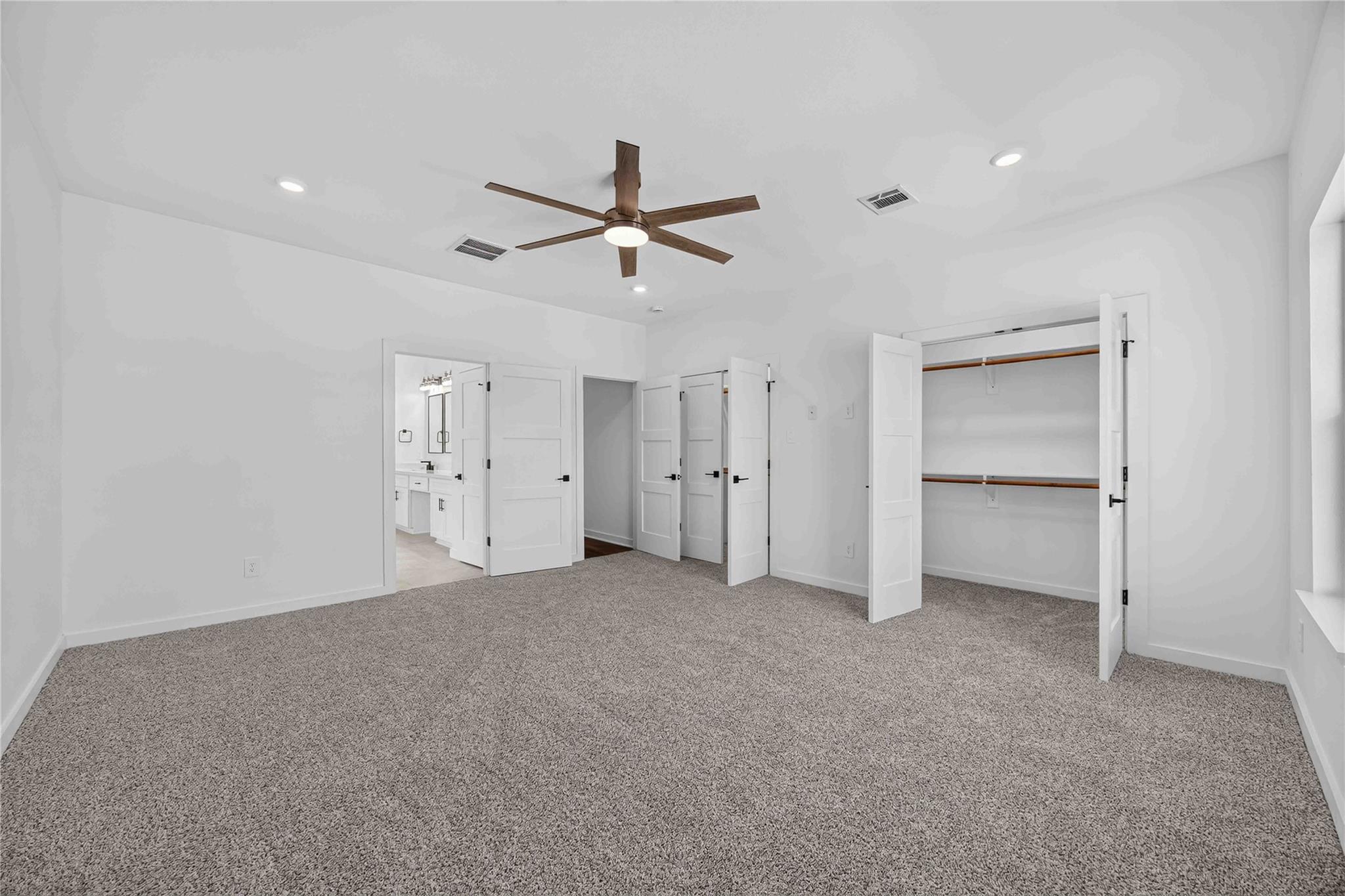 Empty carpeted bedroom with a wood-blade ceiling fan, open closets, and a bathroom doorway.
