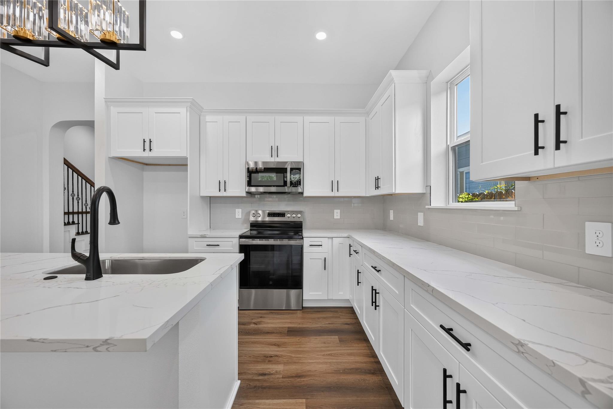 Bright white kitchen with marble-look counters, a large sink island, stainless appliances, and wood flooring.