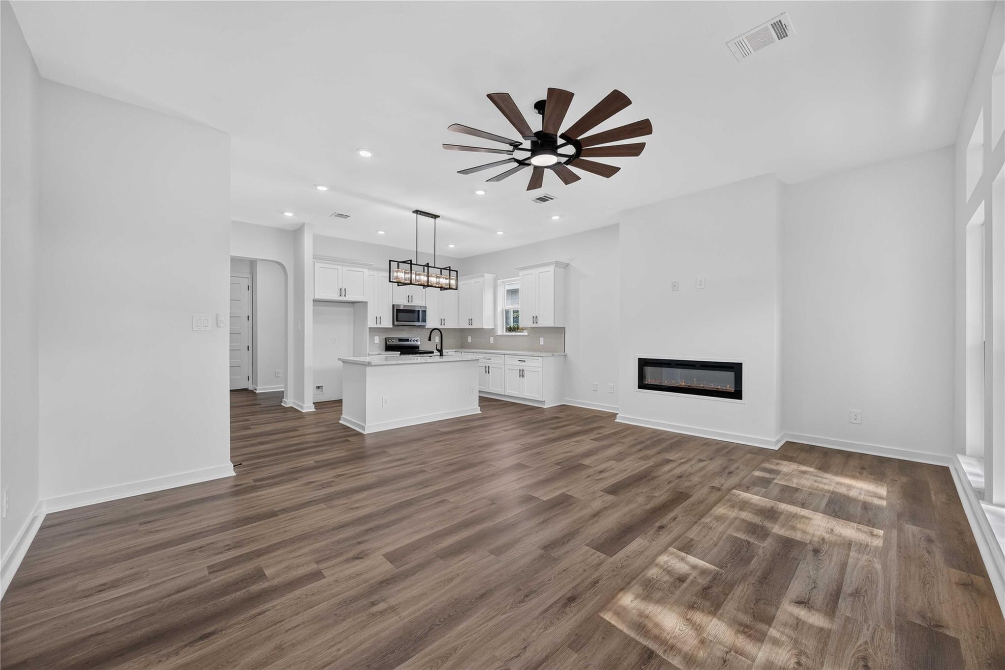 Open-plan living room and kitchen with white cabinets, wood-look floors, a ceiling fan, and linear fireplace.