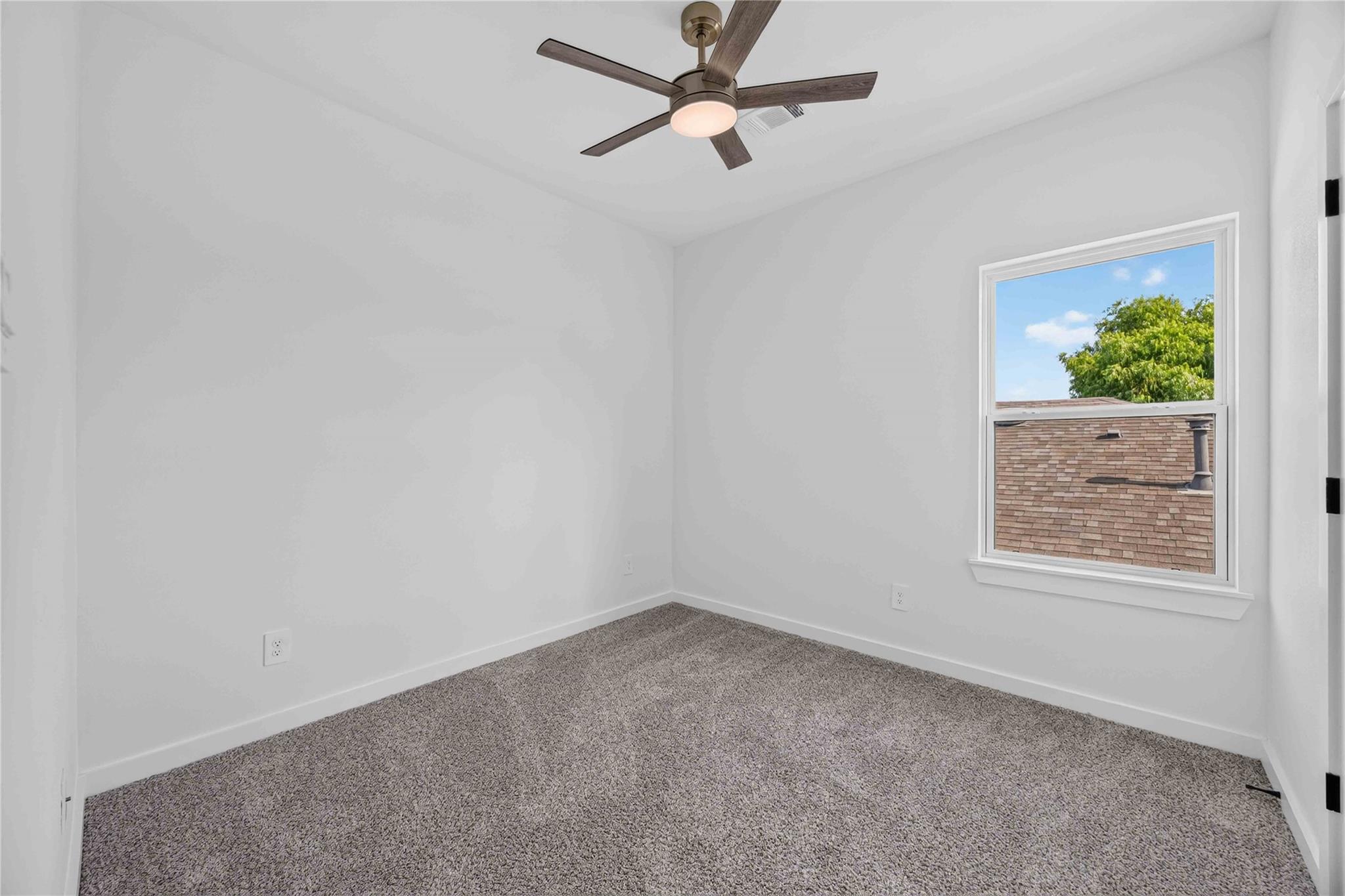 Empty bedroom with gray carpet, ceiling fan, and a window overlooking a leafy tree.
