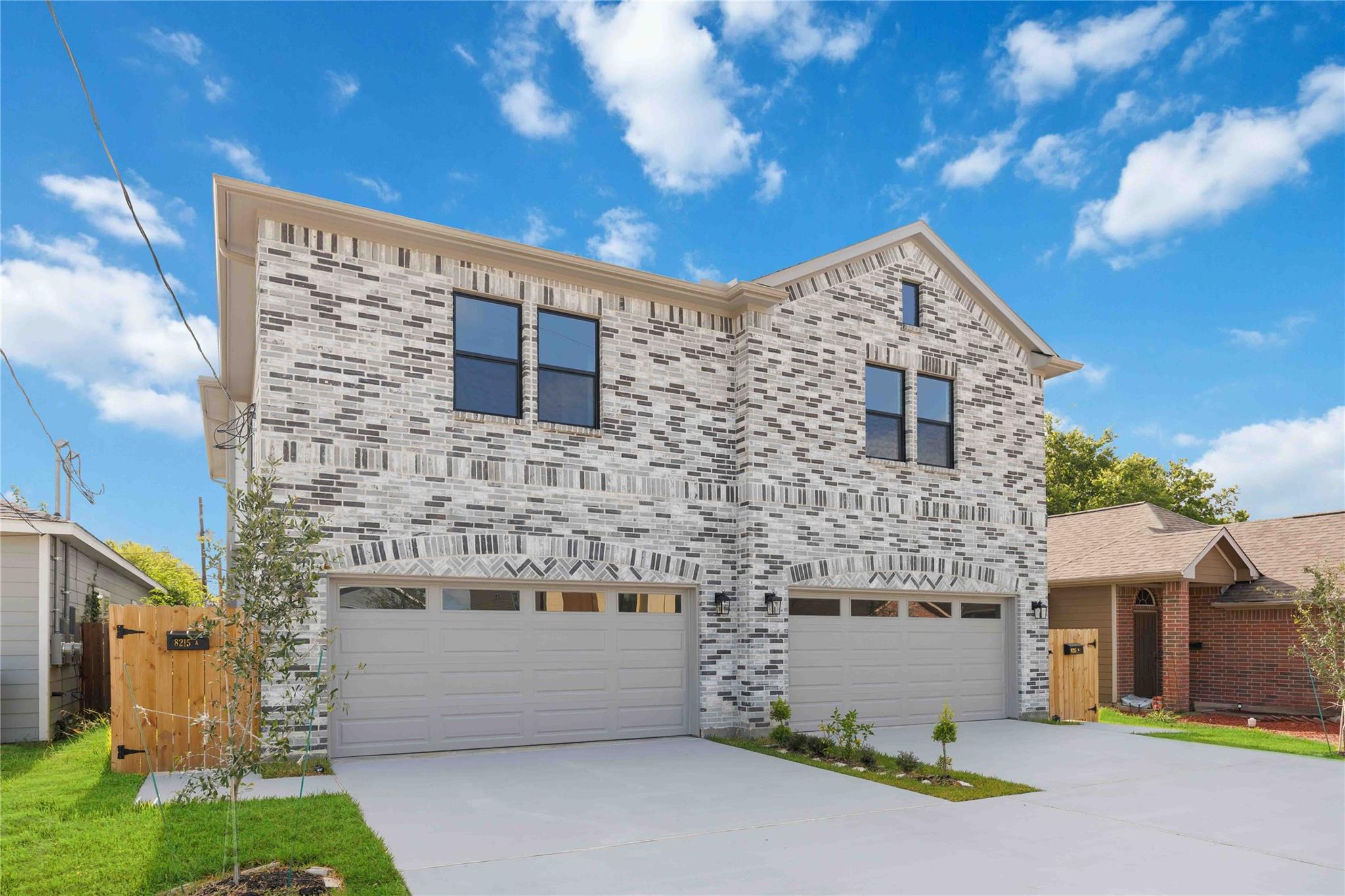 Two-story gray-and-white brick duplex with separate garage doors and driveways, viewed from the front.