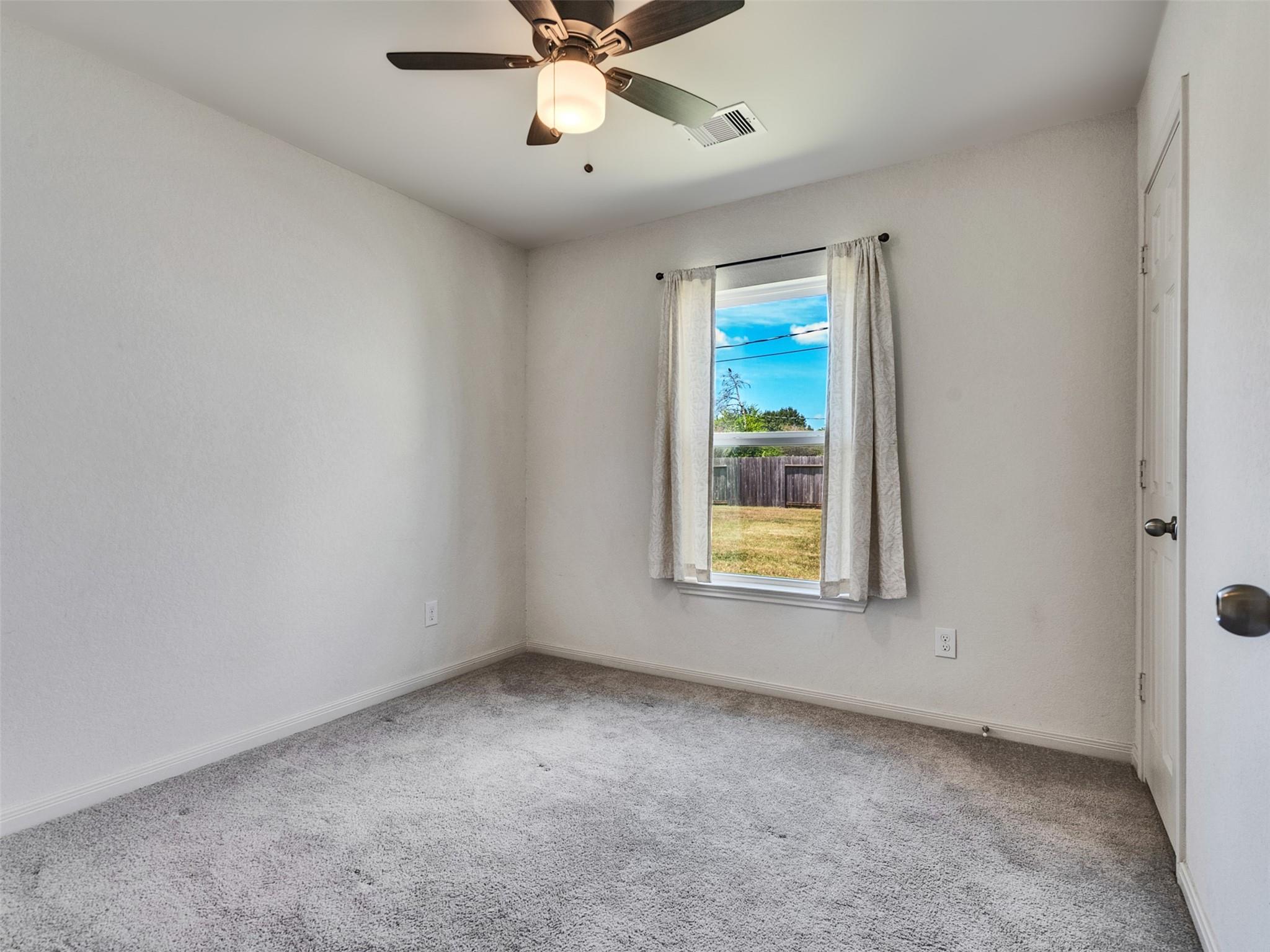 Empty bedroom with gray carpet, ceiling fan, and a curtained window overlooking a fenced yard.