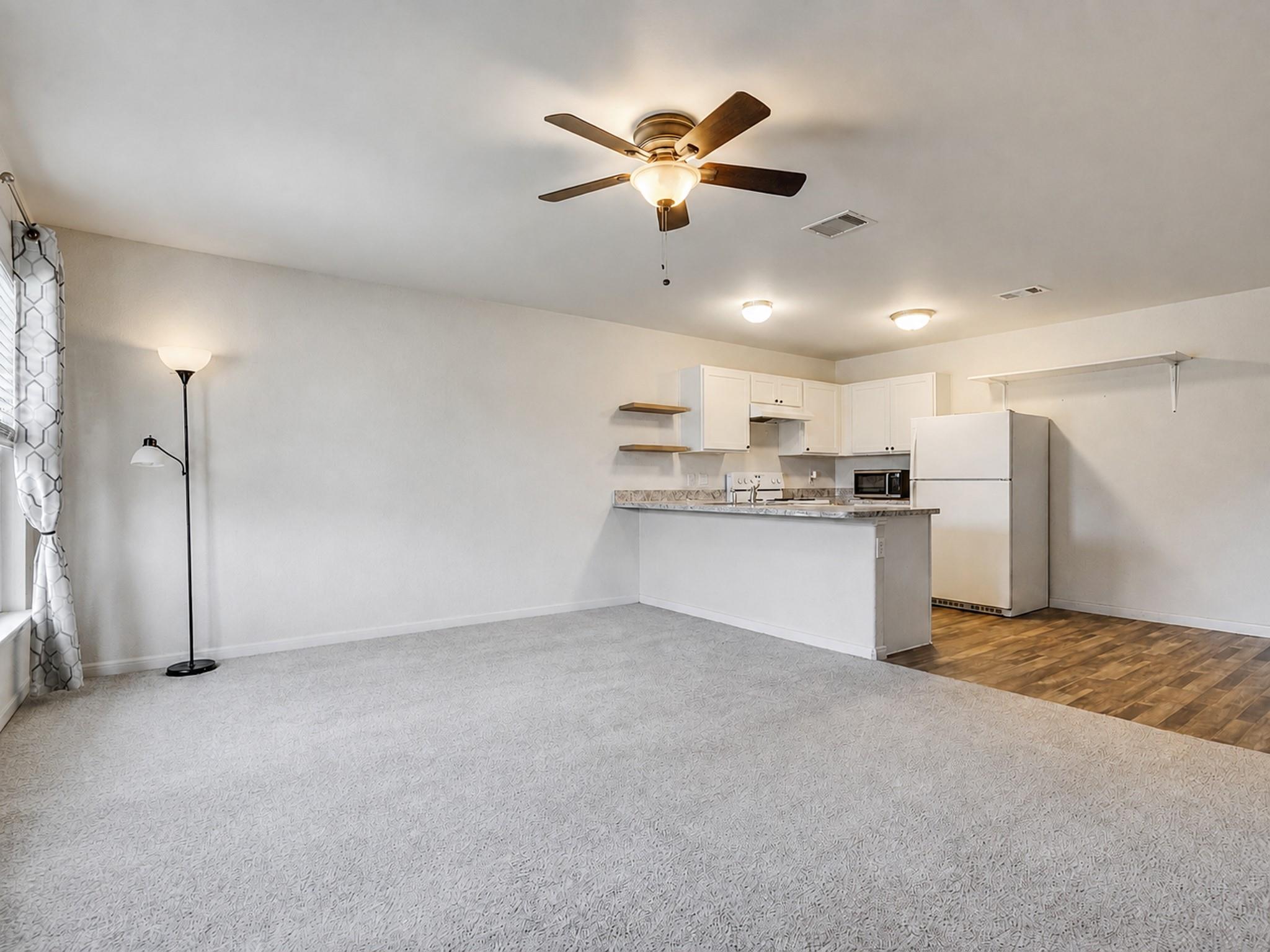 Unfurnished living area with light carpeting, open to a kitchen with white cabinets and a breakfast bar.