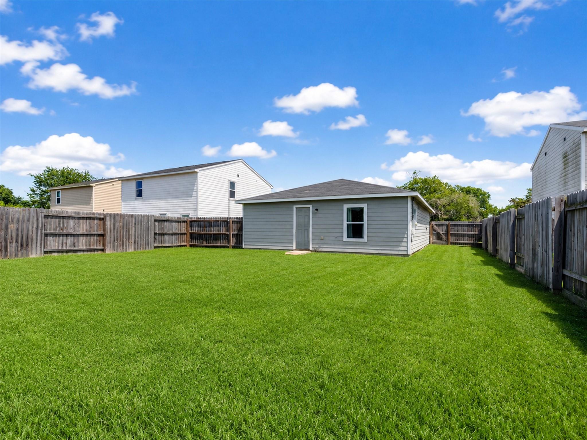 Fenced backyard with a broad green lawn and the rear of a gray single-story house.