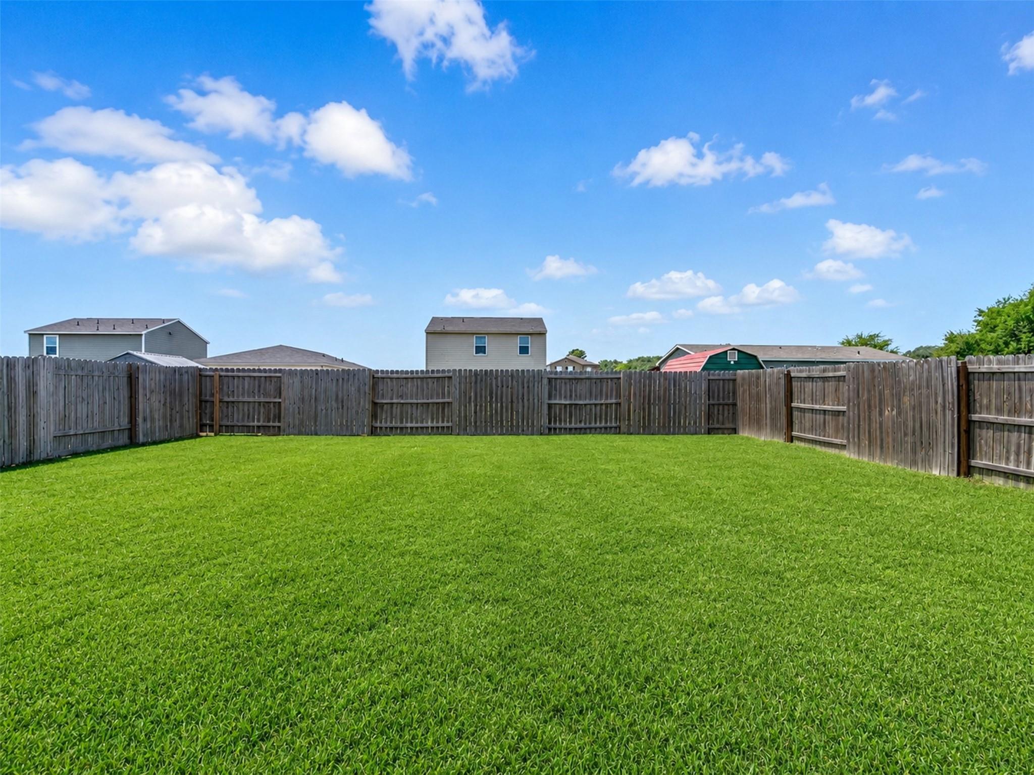 Fenced backyard with a broad green lawn and neighboring houses beyond the fence.