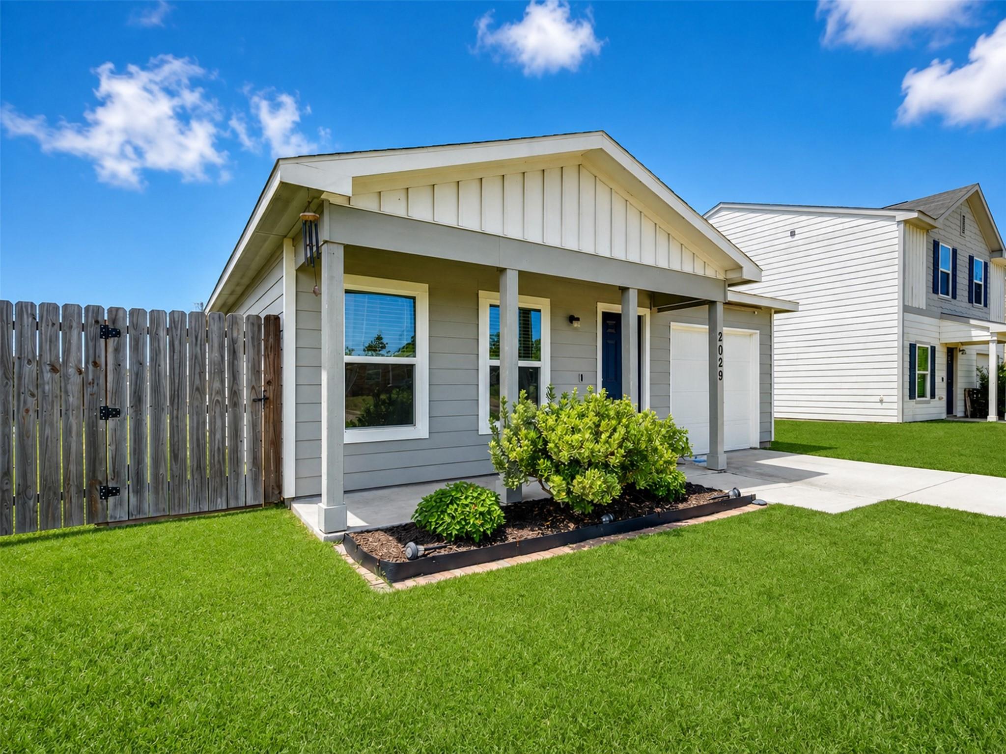 Gray single-story home with a covered porch, white garage, landscaped shrubs, and a fenced green lawn.