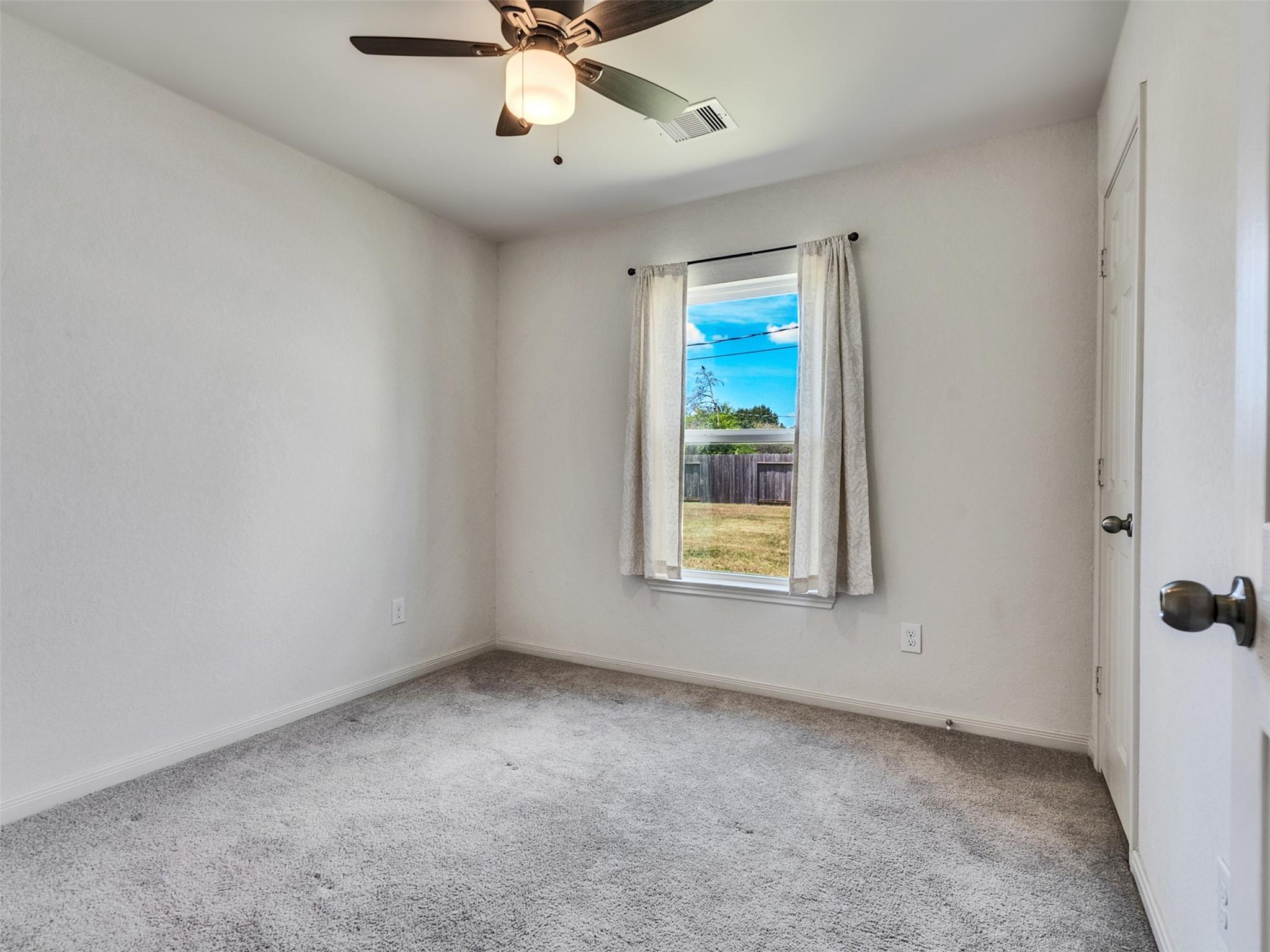 Empty bedroom with gray carpet, a ceiling fan, and a curtained window overlooking a fenced yard.