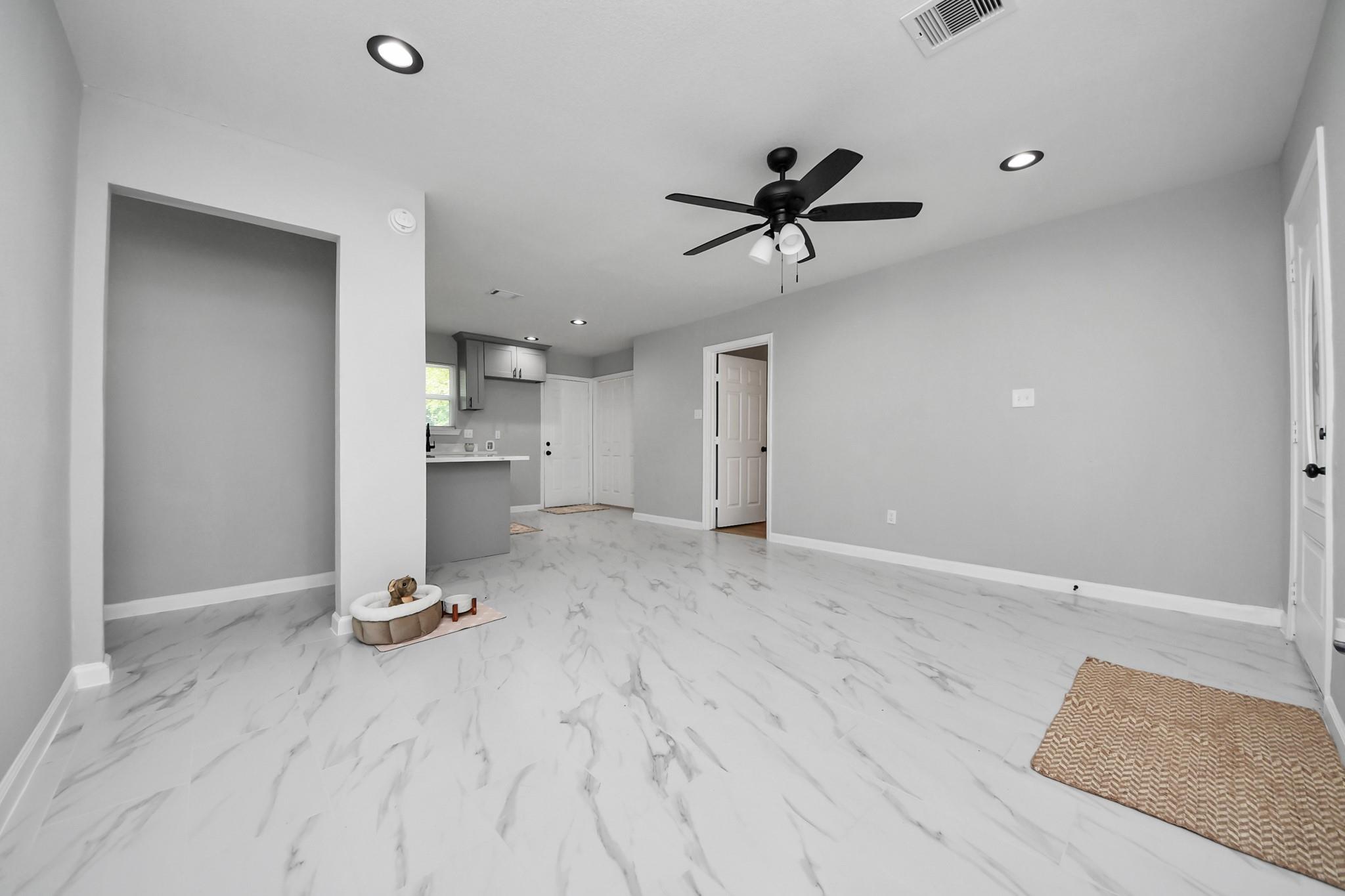 Open living area with marble-look tile floors, a black ceiling fan, and a small kitchen at the back.
