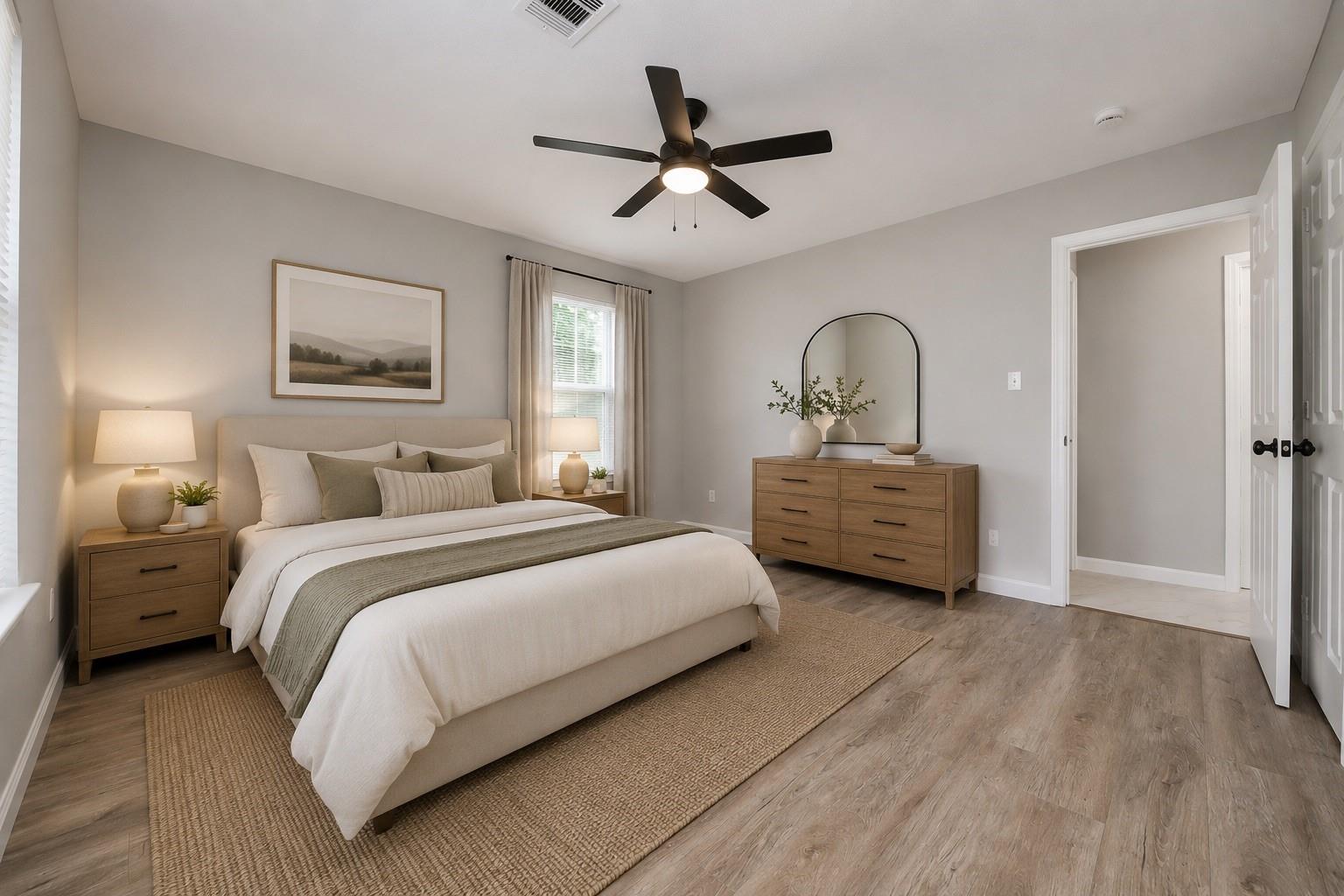 Staged bedroom with a neutral bed, wood furnishings, arched mirror, and ceiling fan.