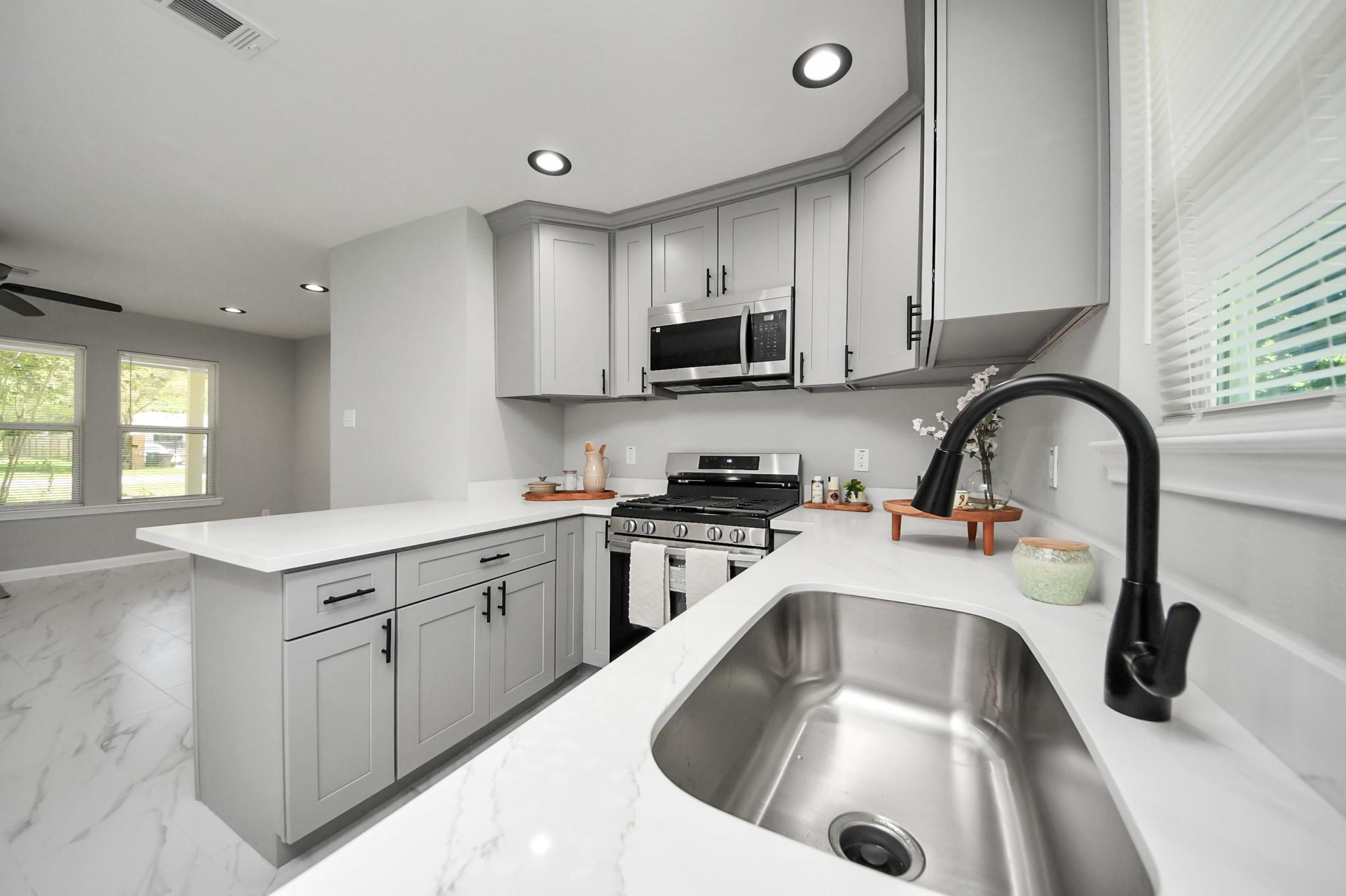 Remodeled kitchen with gray cabinets, white counters, stainless appliances, and a large sink with black faucet.