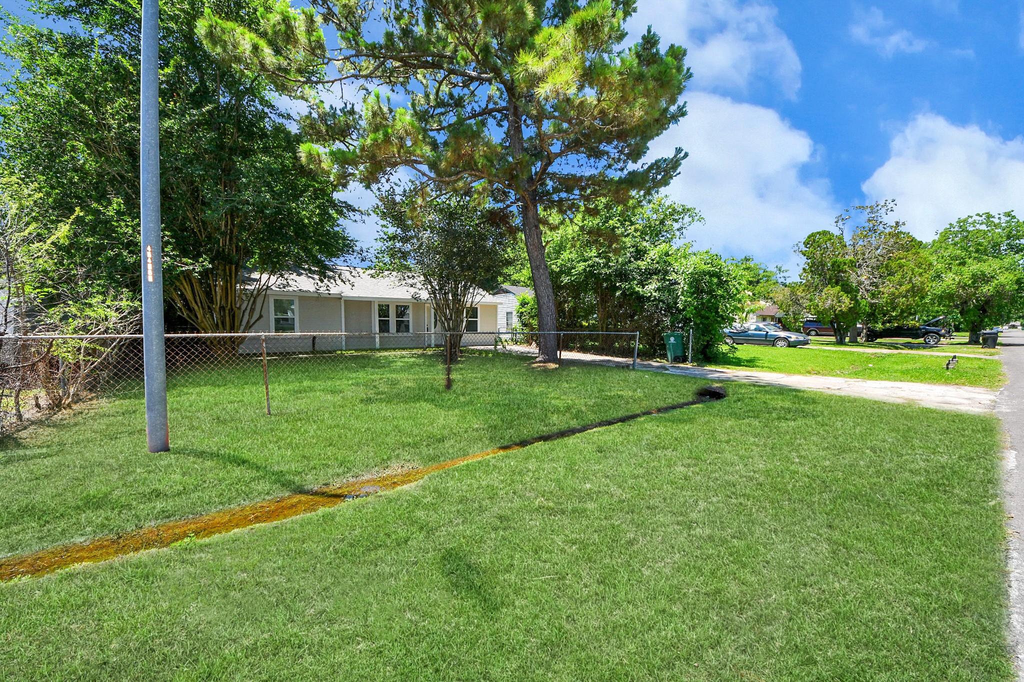 Single-story home behind a chain-link fence and broad grassy yard, shaded by mature trees.