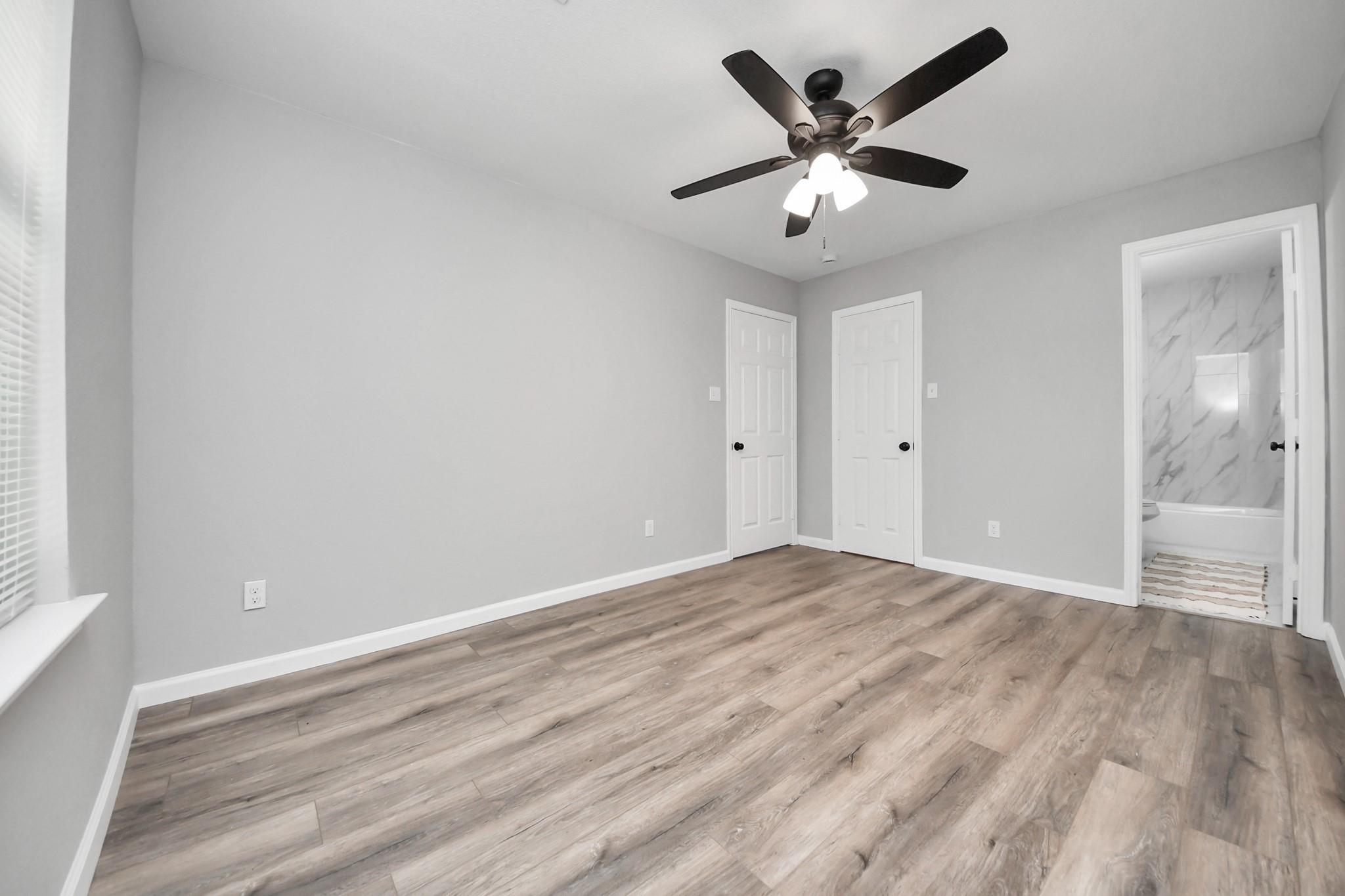 Empty bedroom with wood-look floors, a ceiling fan, and an open doorway to a bathroom.