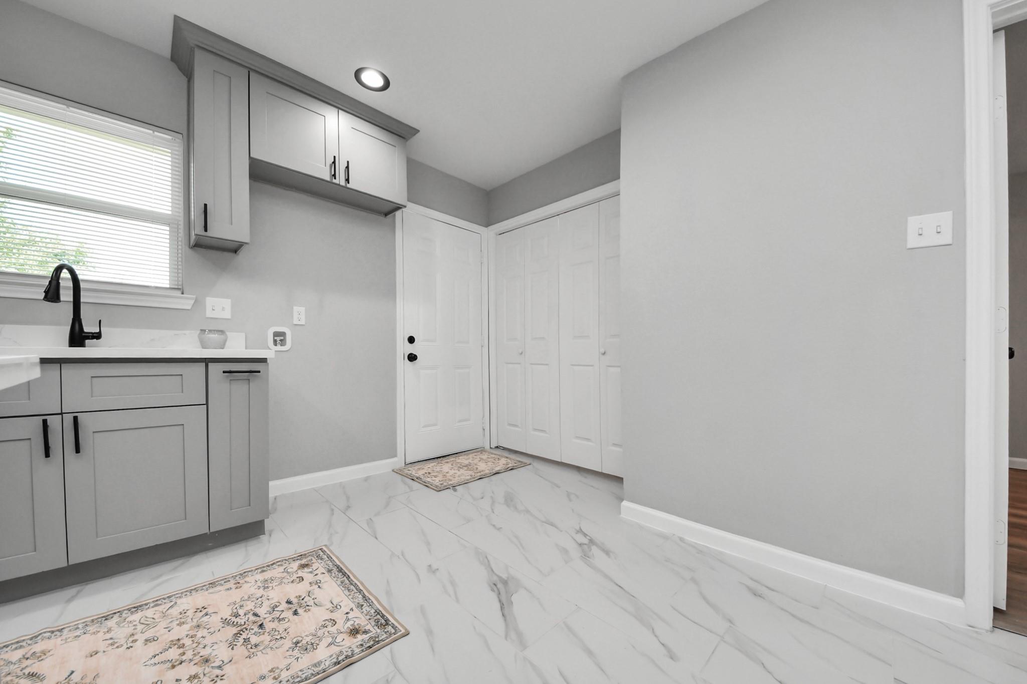 Compact utility area with gray cabinets, a sink beneath a window, closet doors, and marble-look tile flooring.