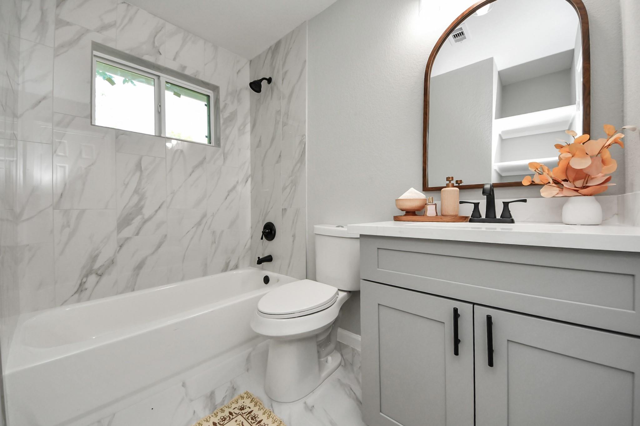 Bathroom with marble-look tub surround, gray vanity, black fixtures, and an arched mirror.
