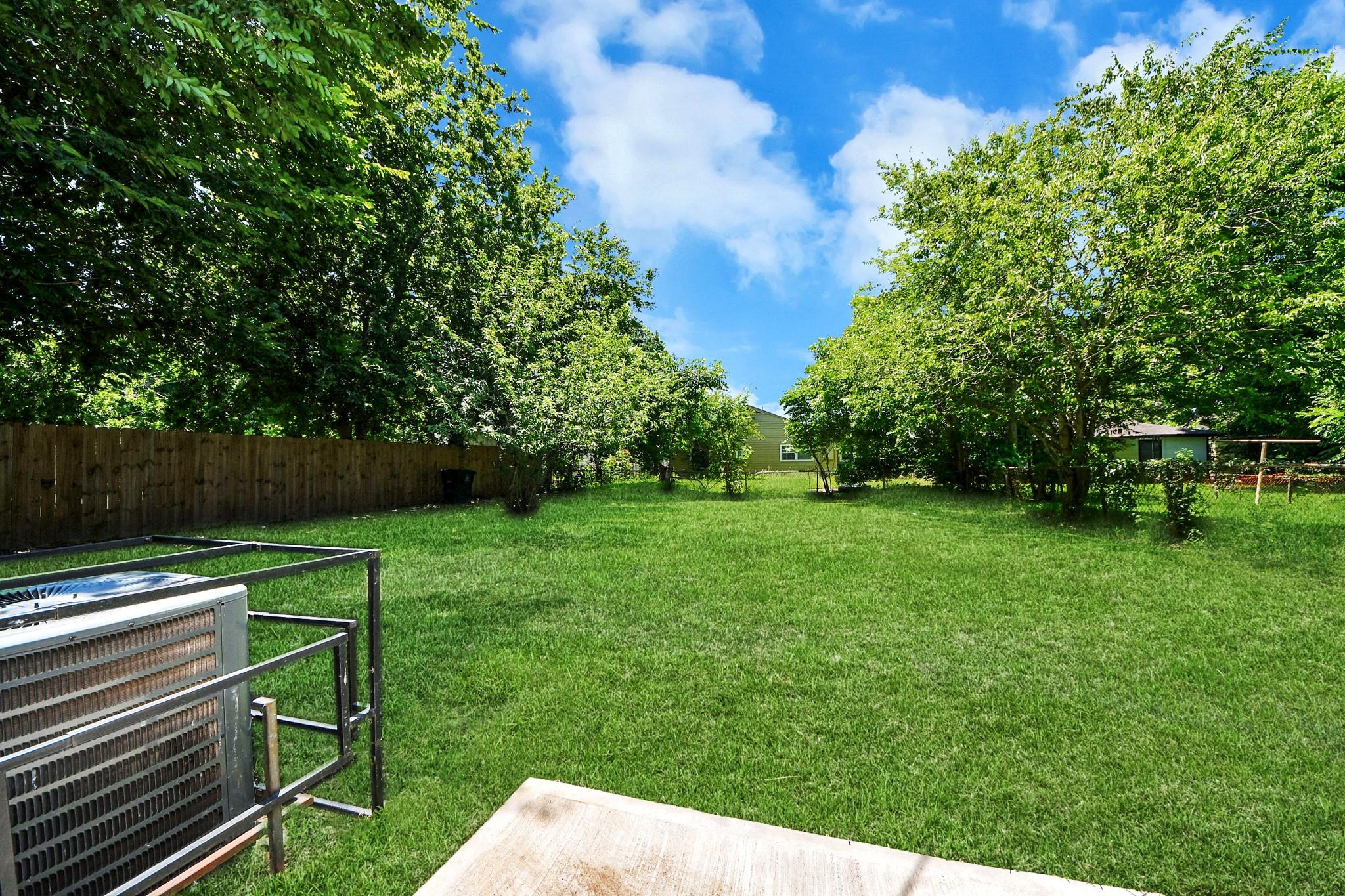 Spacious grassy backyard with mature trees, a wooden fence, and a concrete patio.