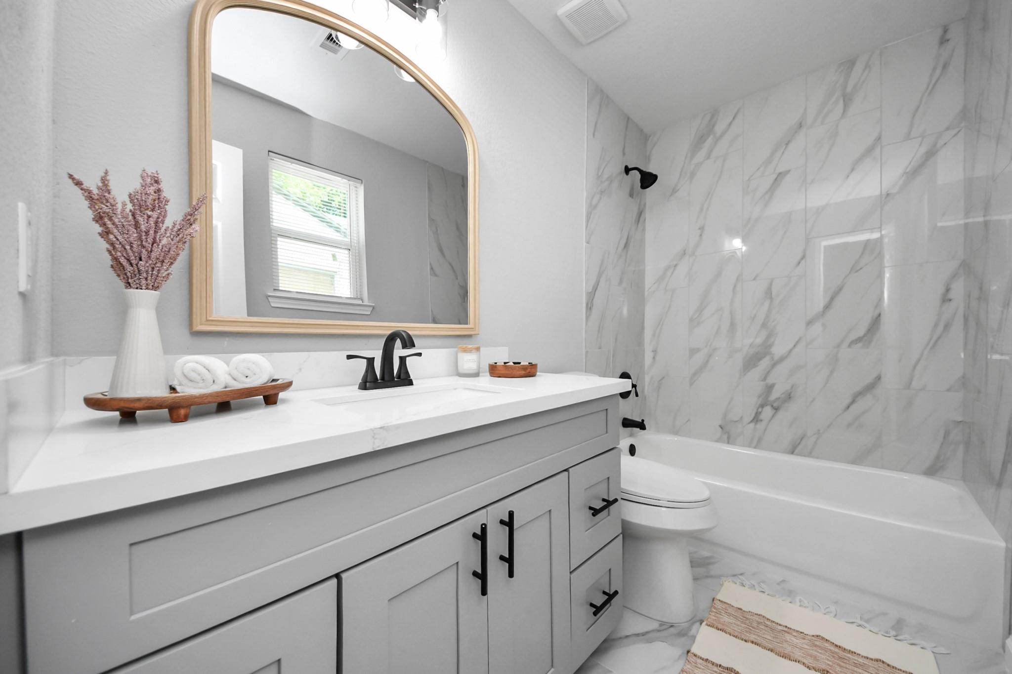 Staged bathroom with a gray vanity, arched gold-framed mirror, and marble-look tub surround.