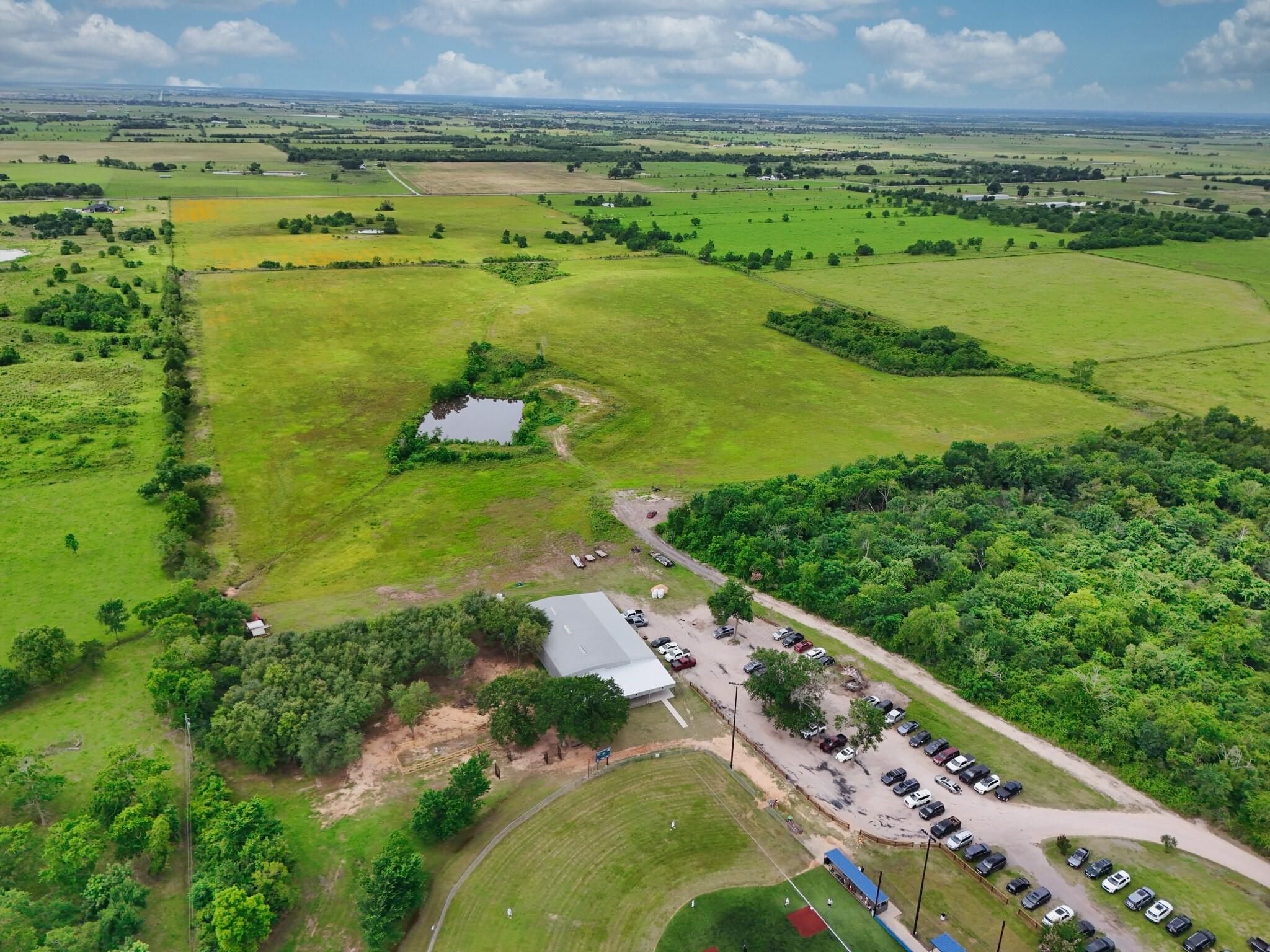 Aerial view of open fields, a pond, and a building with a large parking area.