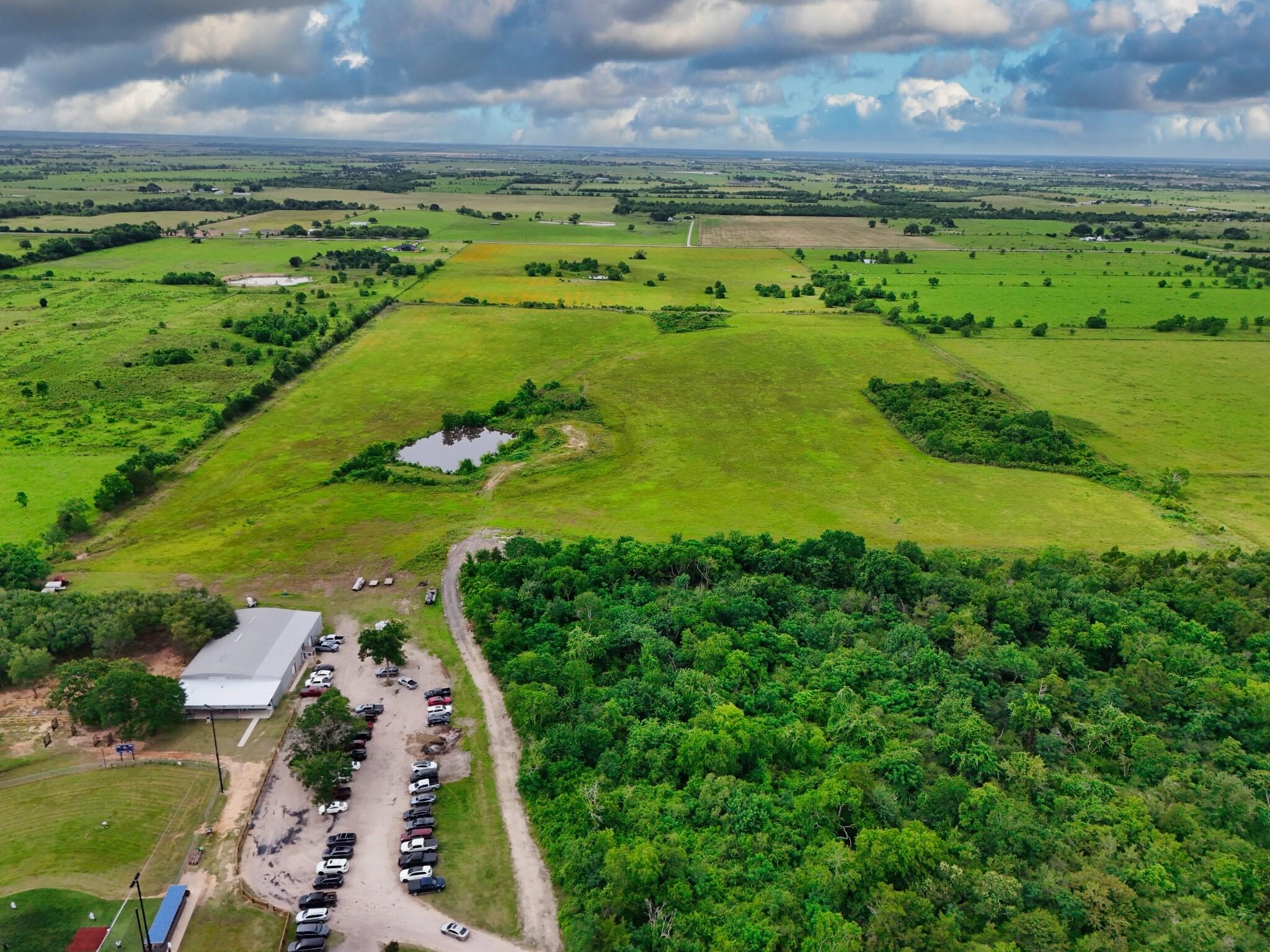 Aerial view of expansive green acreage with a pond, wooded area, and a roadside building.