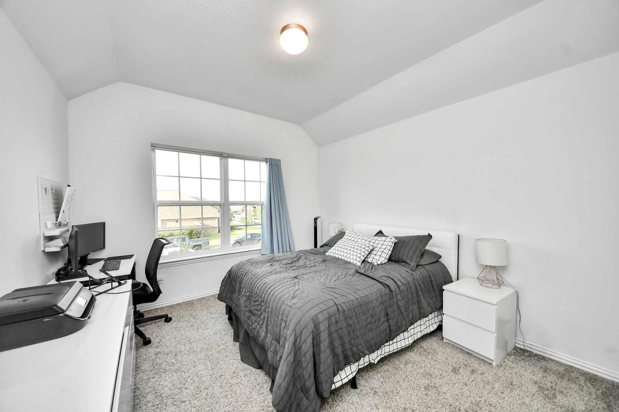 Furnished bedroom with gray bedding, vaulted ceiling, large window, and desk with computer equipment.