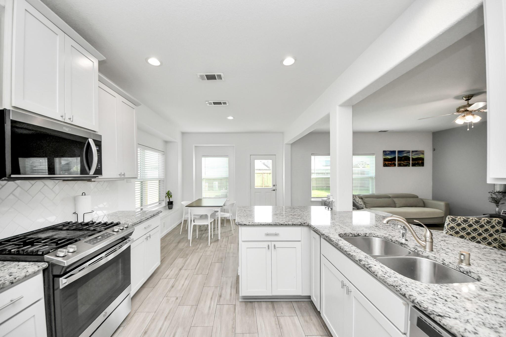 Open-plan kitchen with white cabinets, a granite island and double sink, opening to a dining nook and living room.