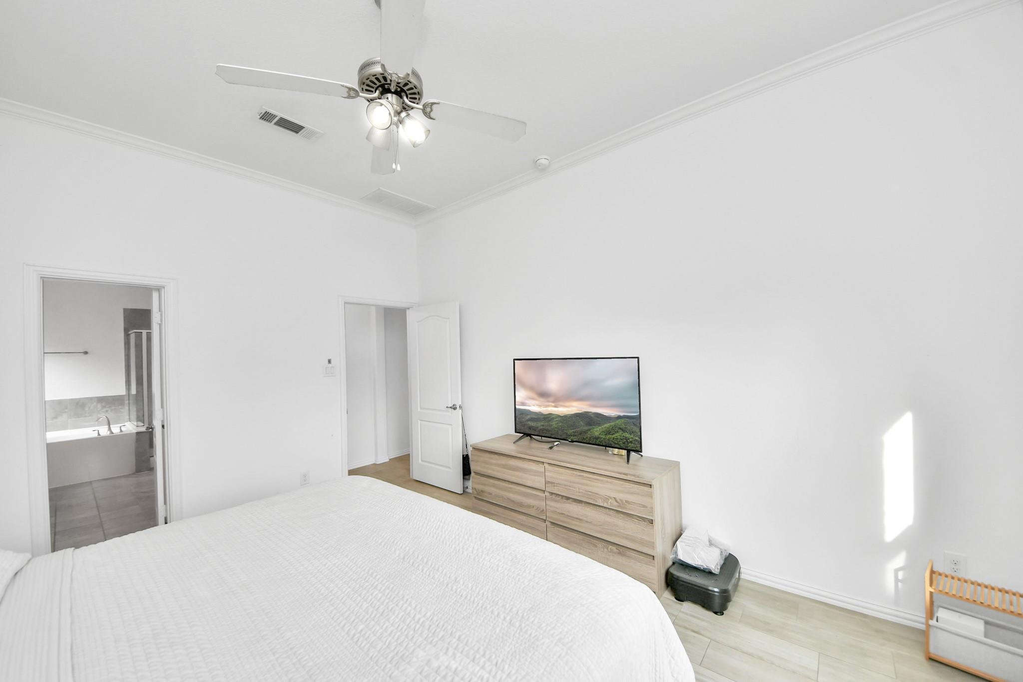 Bright bedroom with a white bed, ceiling fan, and television on a light wood dresser.