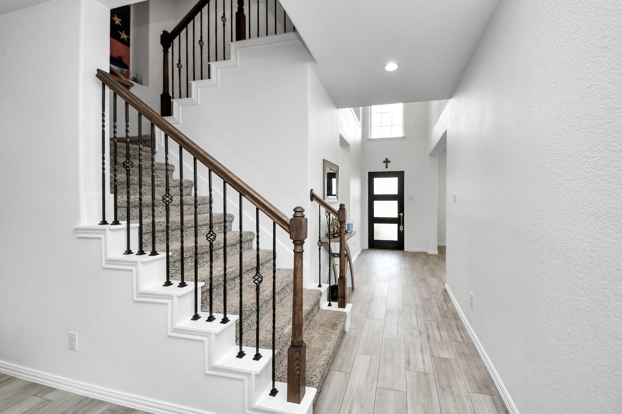Entry foyer with carpeted staircase, dark wood railing, light tile floor, and black front door with frosted glass.
