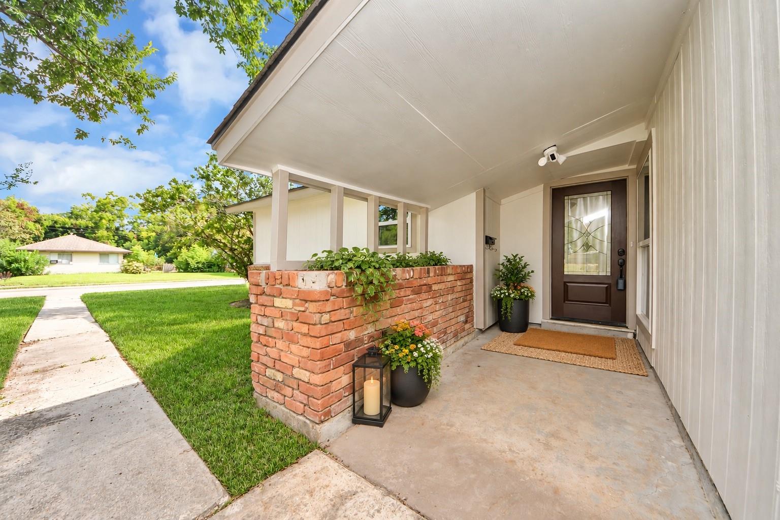 Covered front porch with a dark glass-paneled door, brick planter wall, and potted flowers.