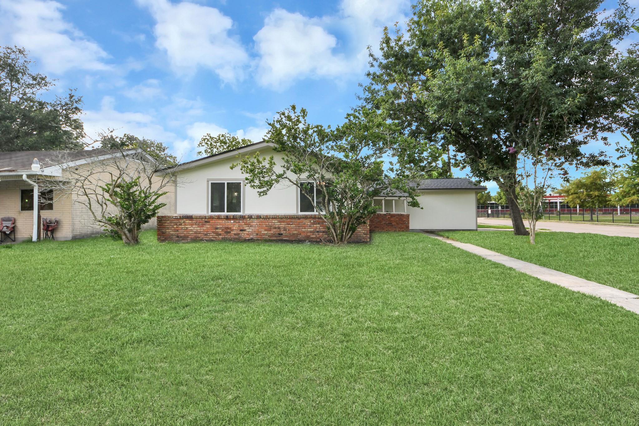 Single-story home with a red brick lower facade, broad lawn, mature trees, and a concrete walkway.