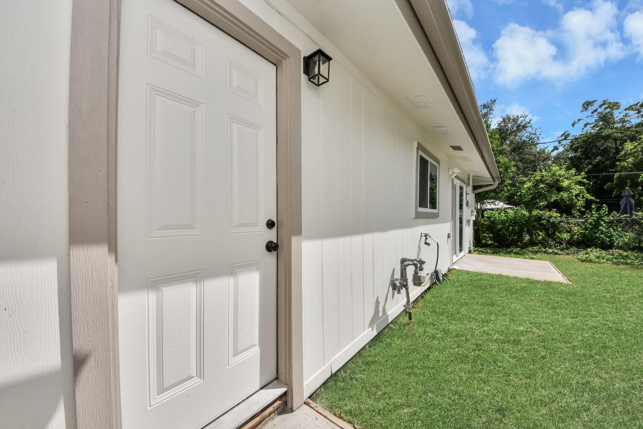 White-sided home’s side entrance with a paneled door, wall lantern, and lawn.