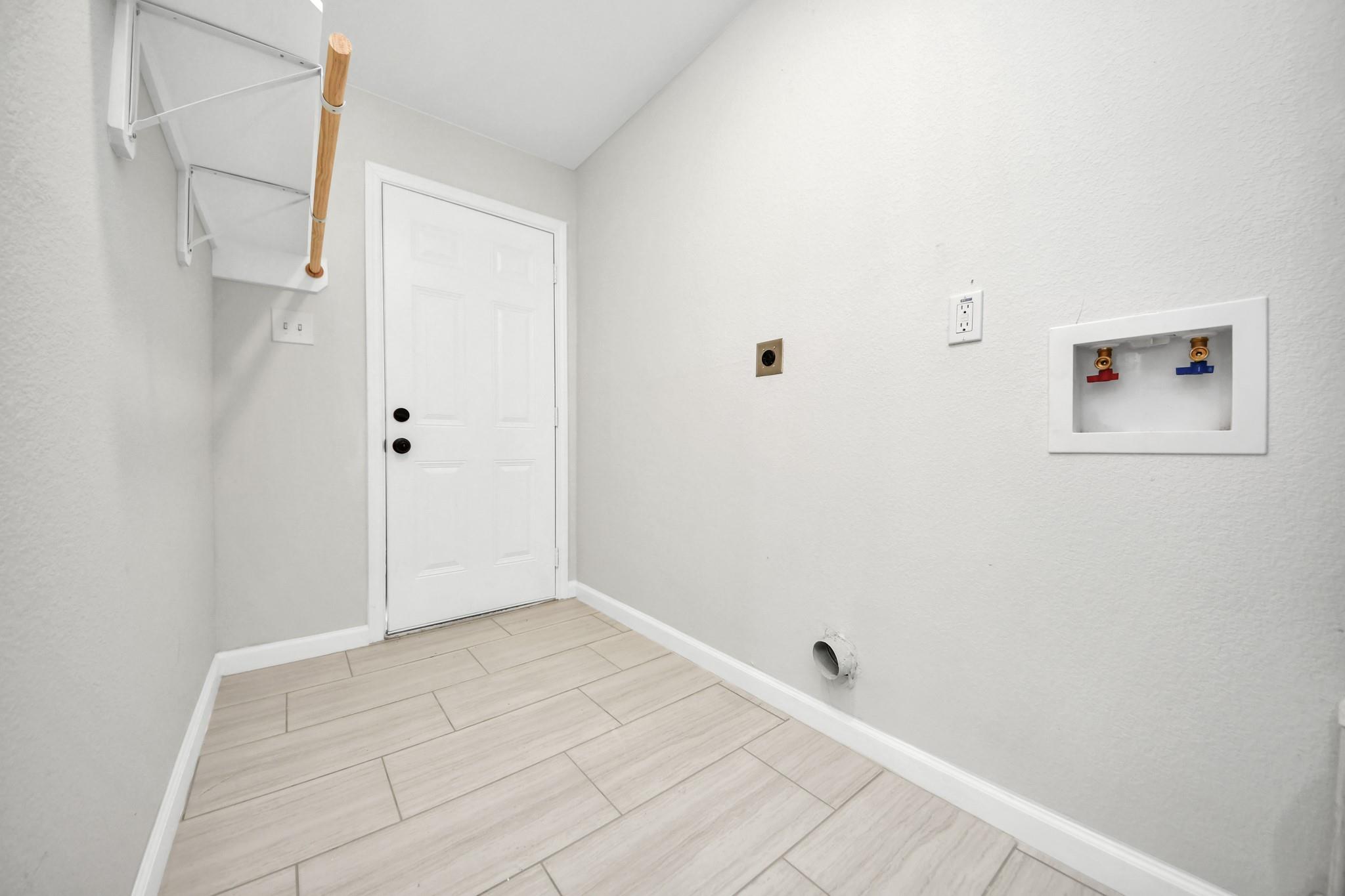 Empty laundry room with pale tile floors, wall shelves, and recessed washer hookups.