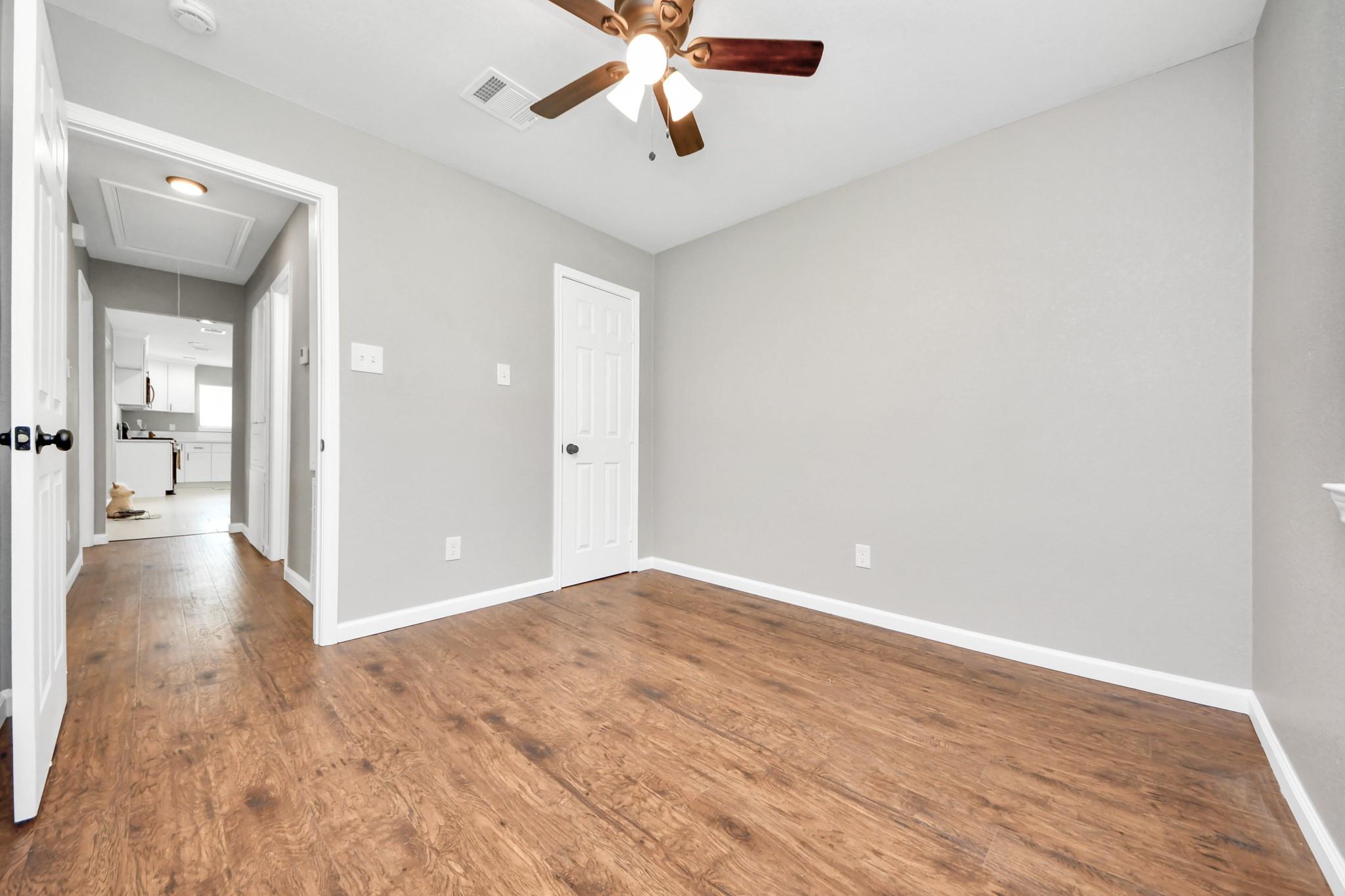 Empty room with wood-look floors and ceiling fan, opening to a hallway and kitchen.