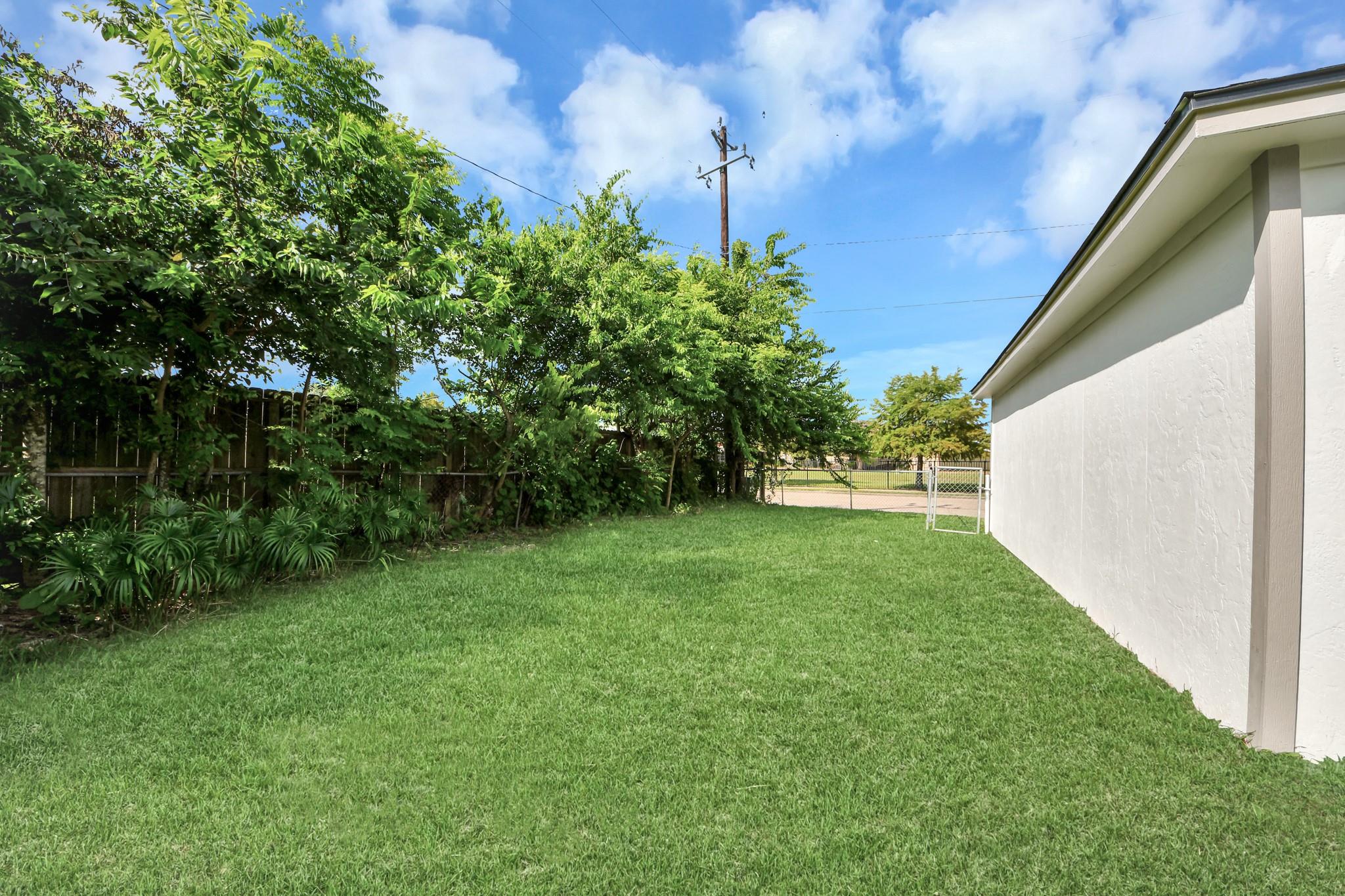 Side yard with a broad green lawn, mature trees along a wooden fence, and a metal gate.