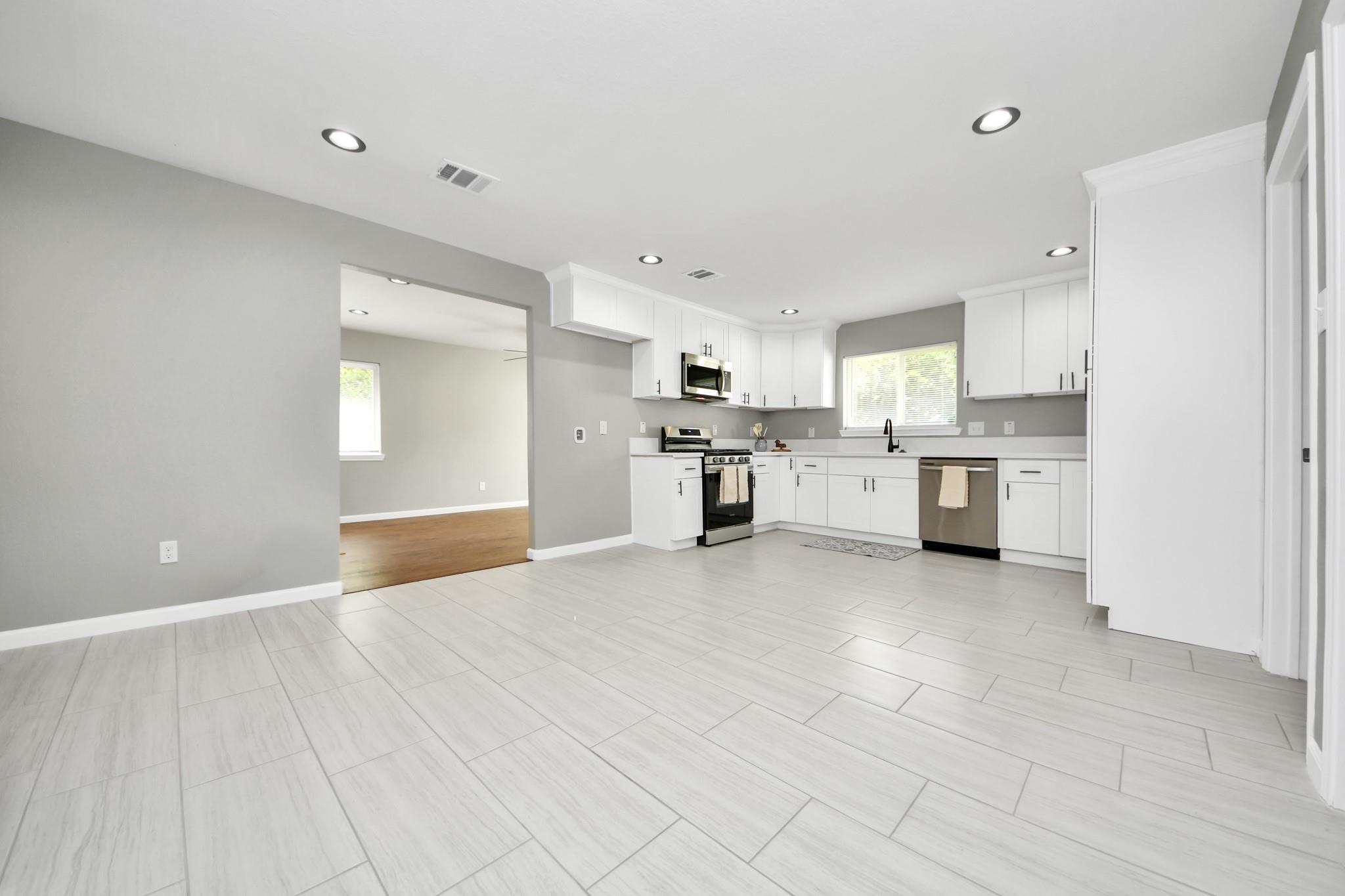 Updated kitchen with white cabinets, stainless steel appliances, and light tile flooring opening to a wood-floored room.