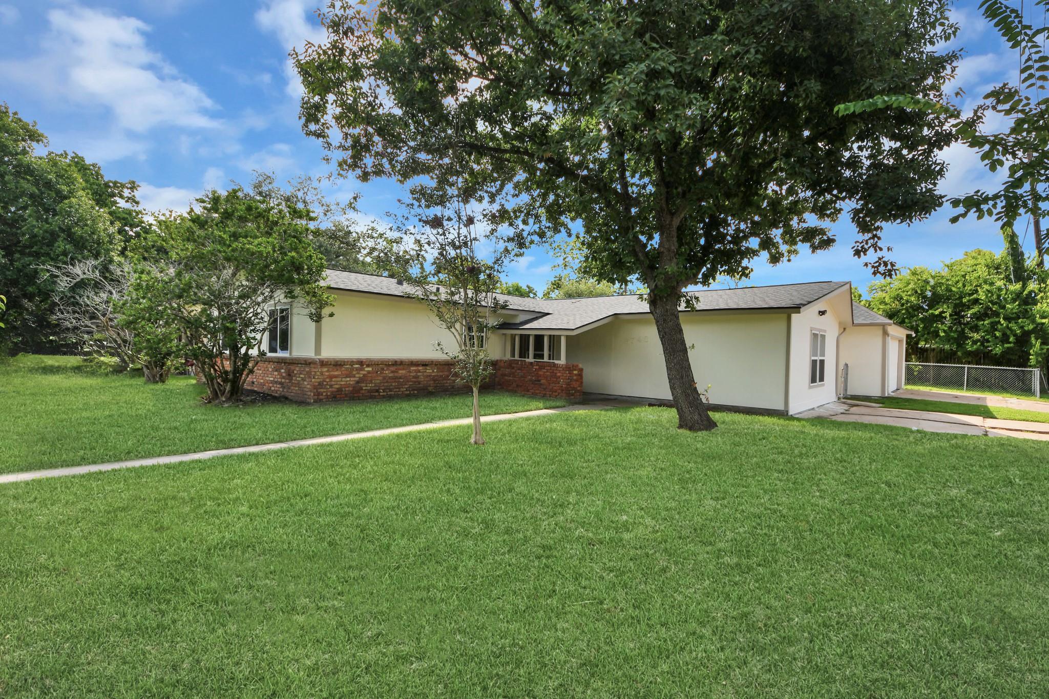 Single-story cream home with a red brick lower facade, mature shade trees, and a broad lawn.