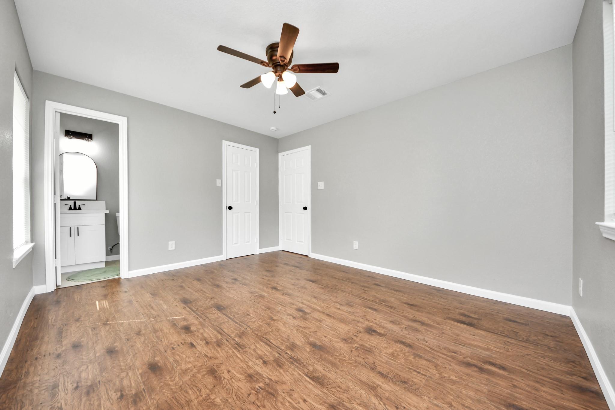 Empty bedroom with wood-look flooring, ceiling fan, and an open doorway to a bathroom.