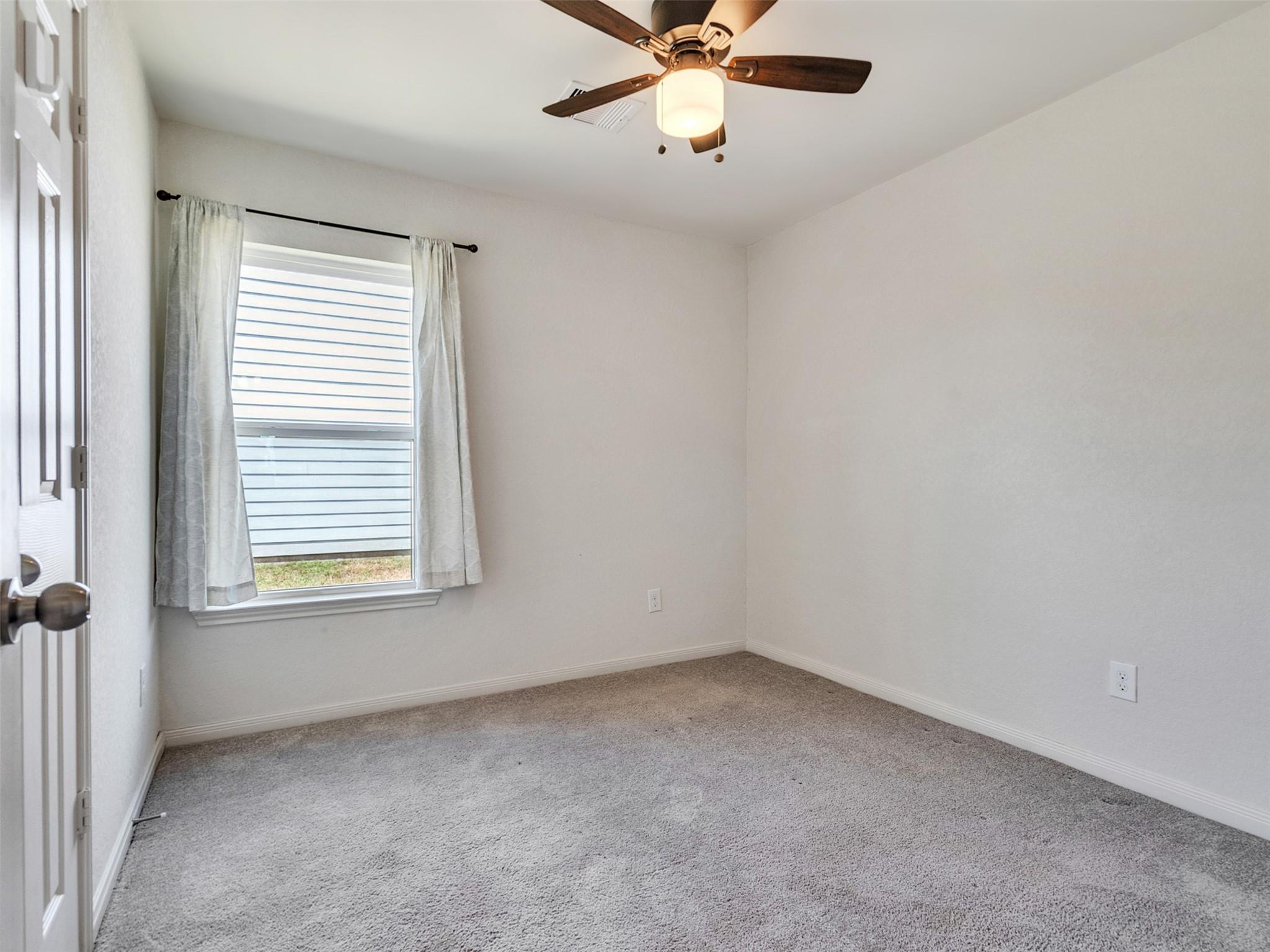 Empty bedroom with gray carpet, a curtained window, and a ceiling fan with a light.