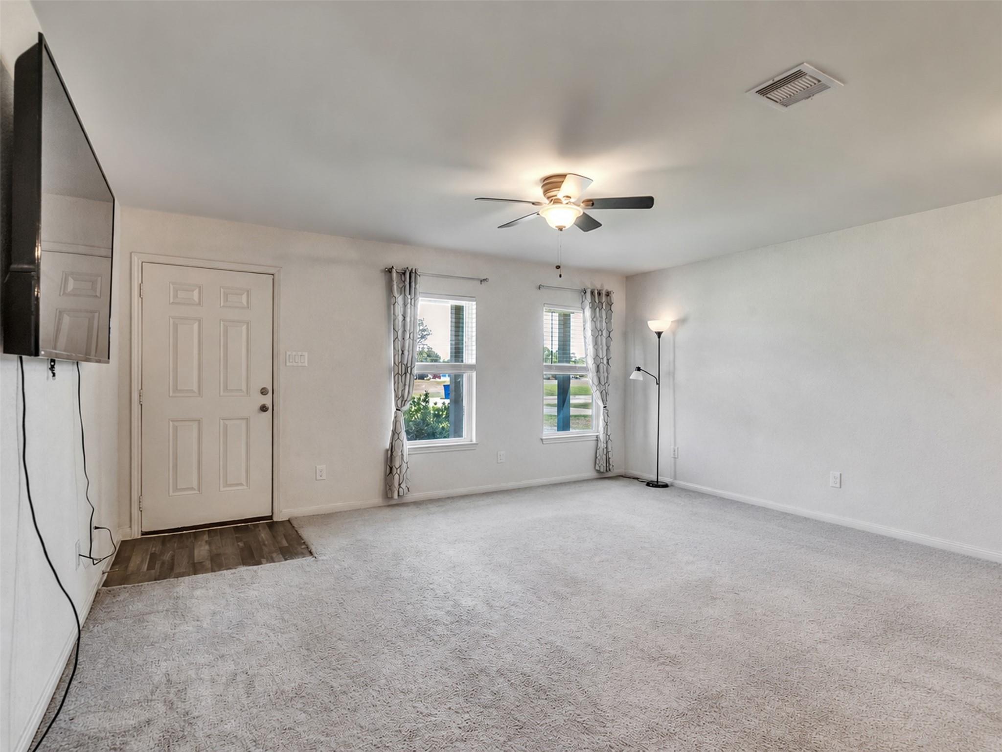 Empty living room with light carpet, ceiling fan, two curtained windows, and a front door.