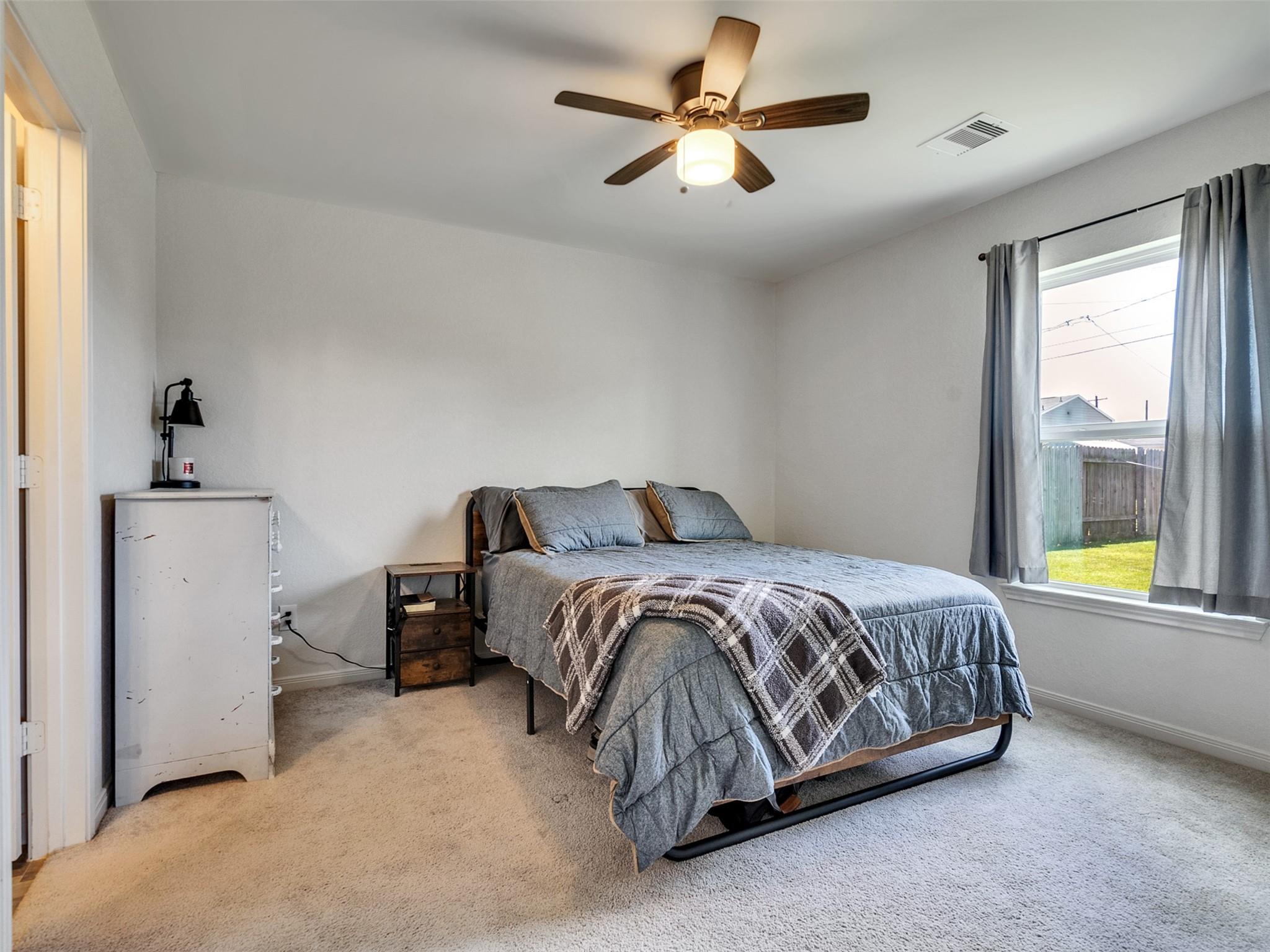 Furnished bedroom with a gray bed, patterned throw, ceiling fan, and window overlooking a fenced yard.