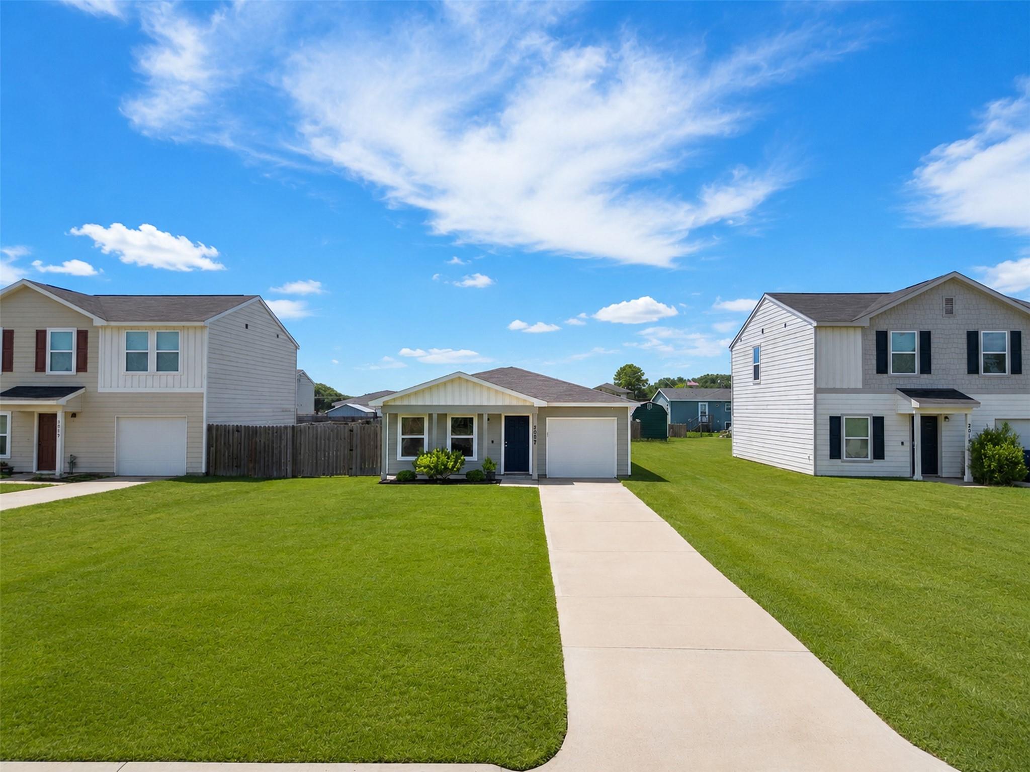 Single-story light-colored house with a blue door, attached garage, and long concrete driveway across a green lawn.