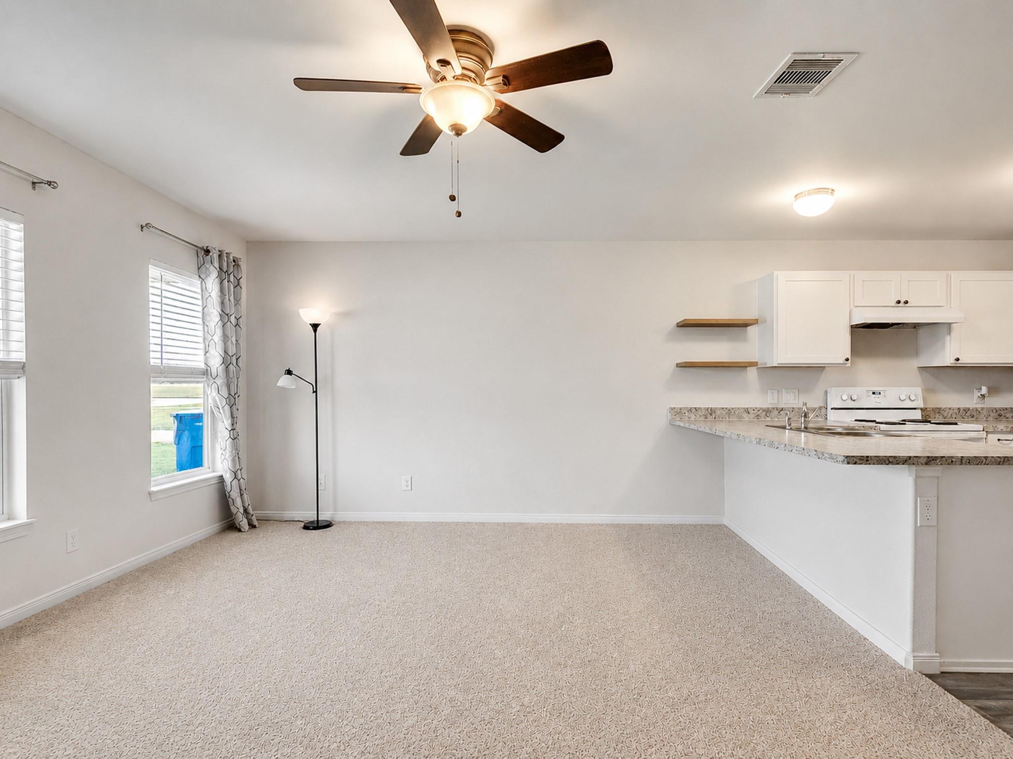 Empty carpeted living area with a ceiling fan and an adjoining kitchen with white cabinets.