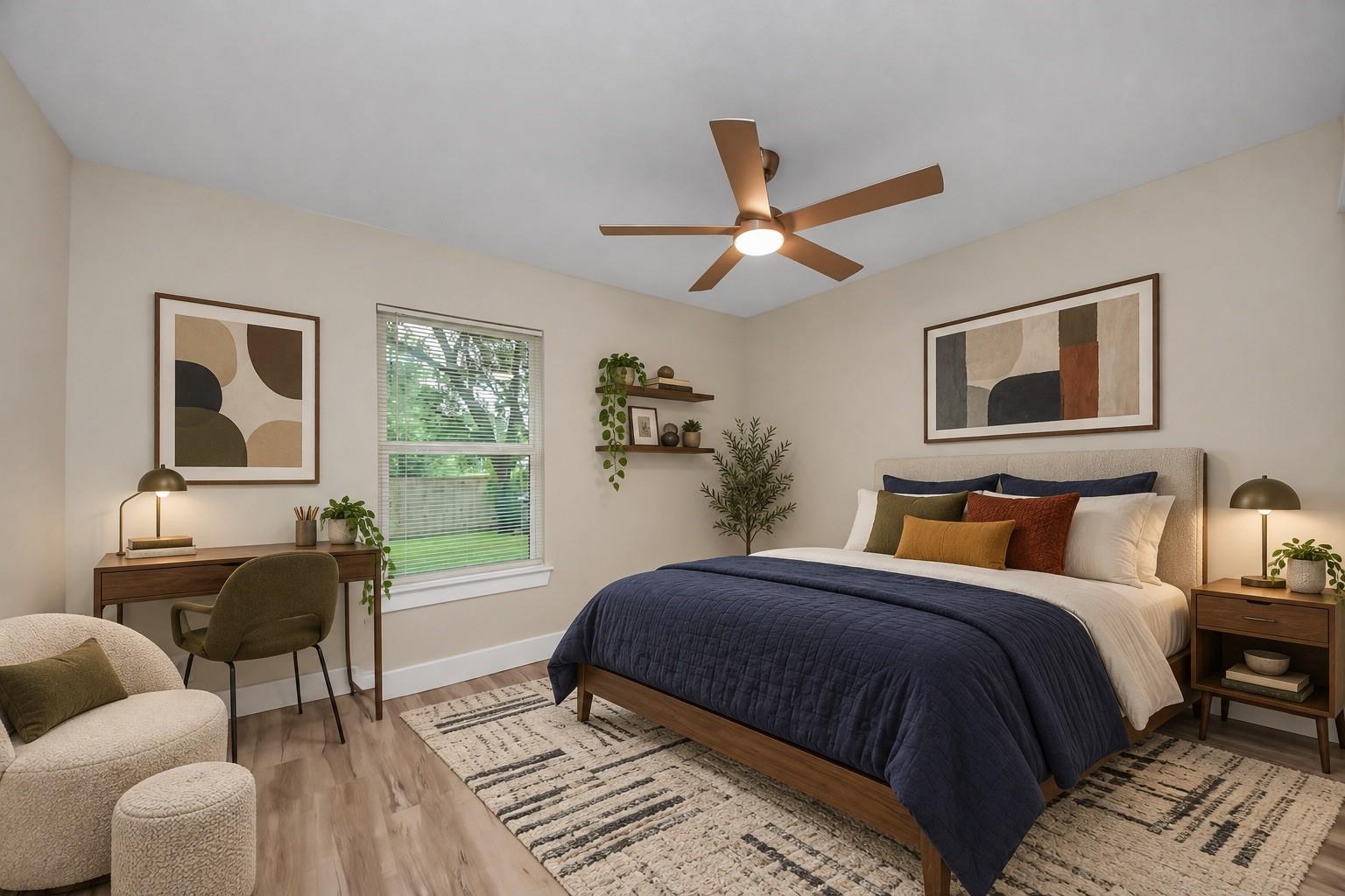 Staged bedroom with a navy-covered bed, ceiling fan, and desk beside a window.