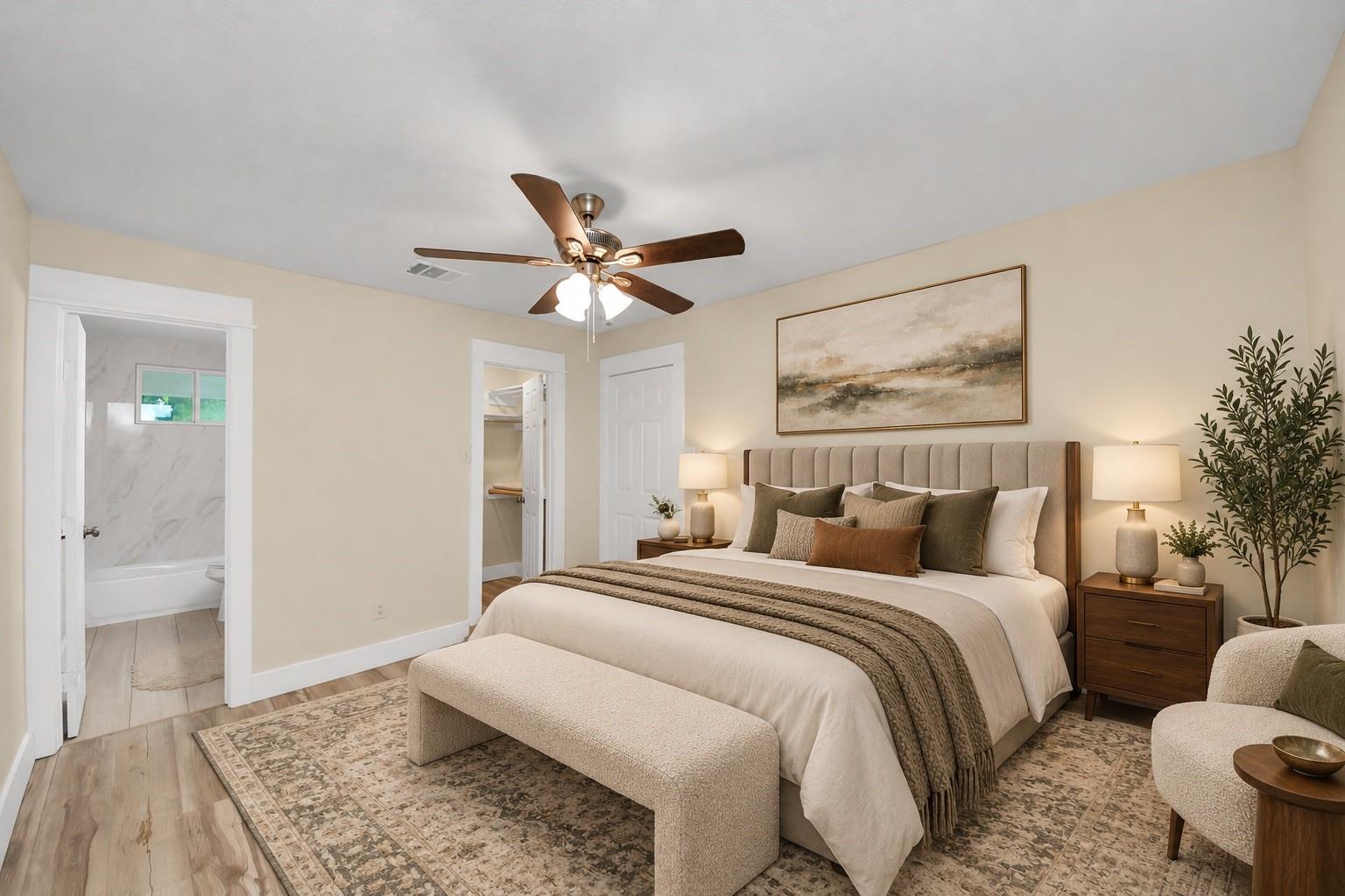 Staged bedroom with an upholstered bed, ceiling fan, patterned rug, and adjoining bathroom doorway.