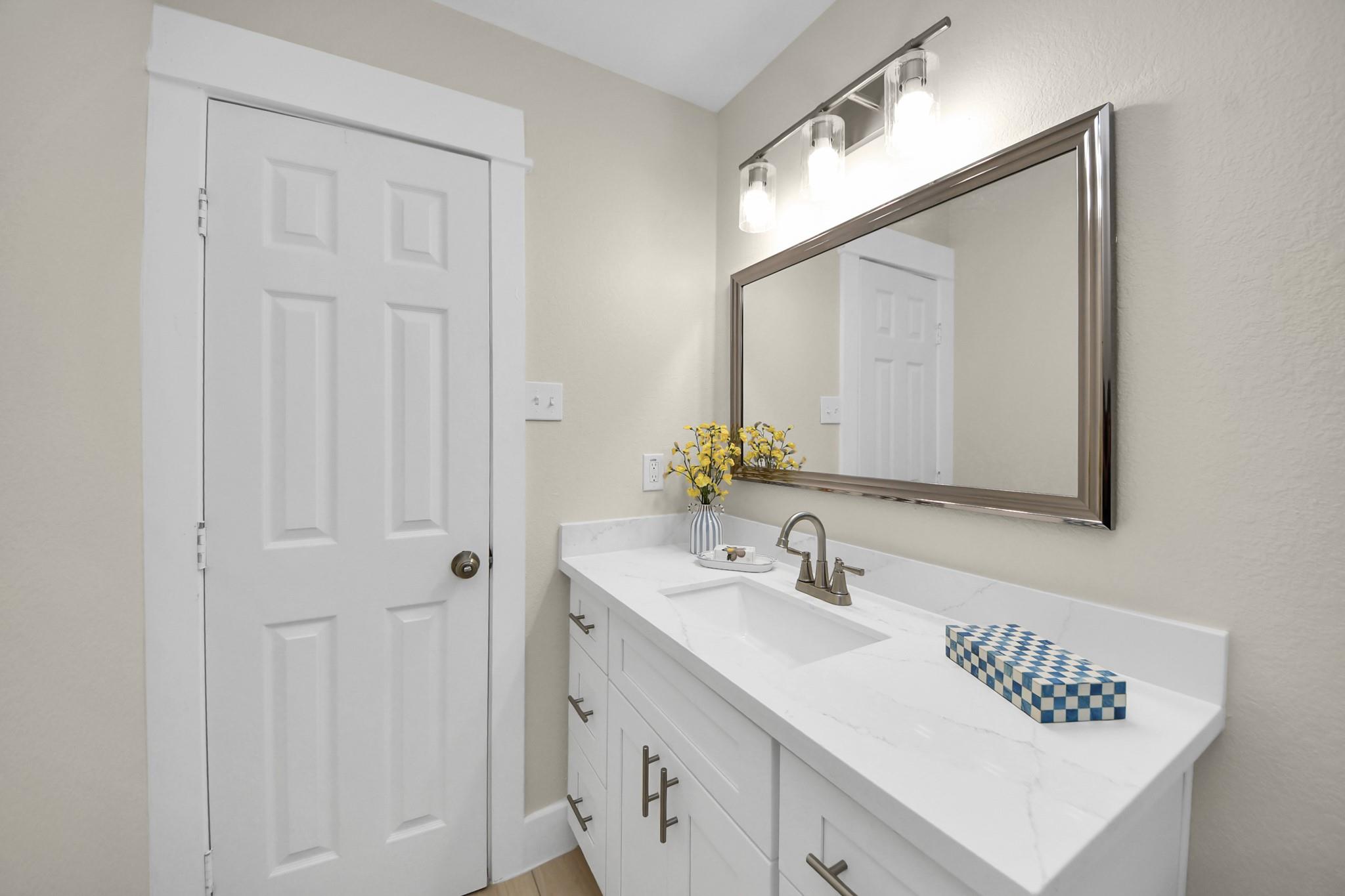 Bathroom vanity with white cabinetry, marble-look counter, framed mirror, and yellow flowers.