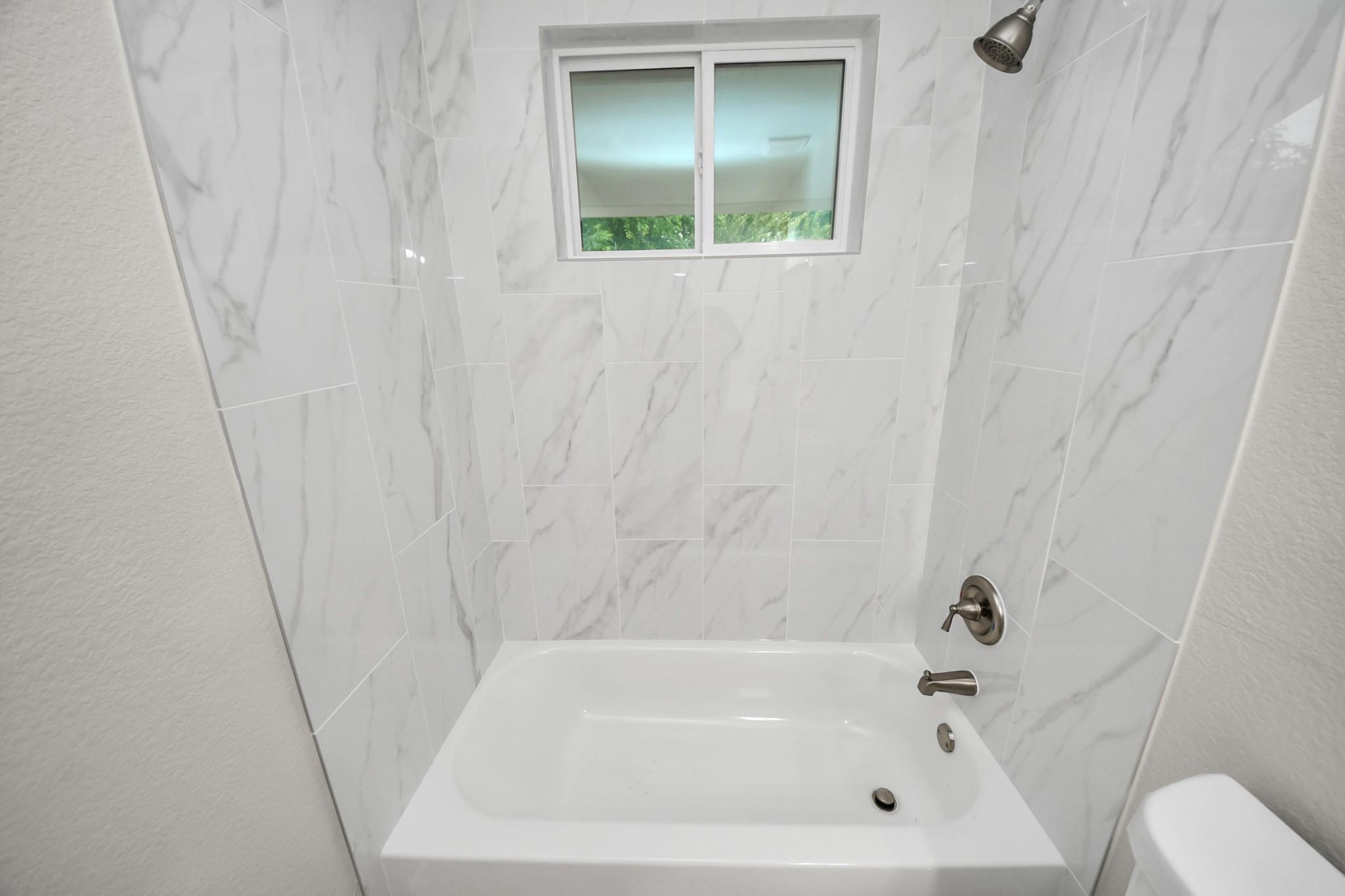 Bathroom bathtub with marble-look tile, brushed metal fixtures, and a small window above.