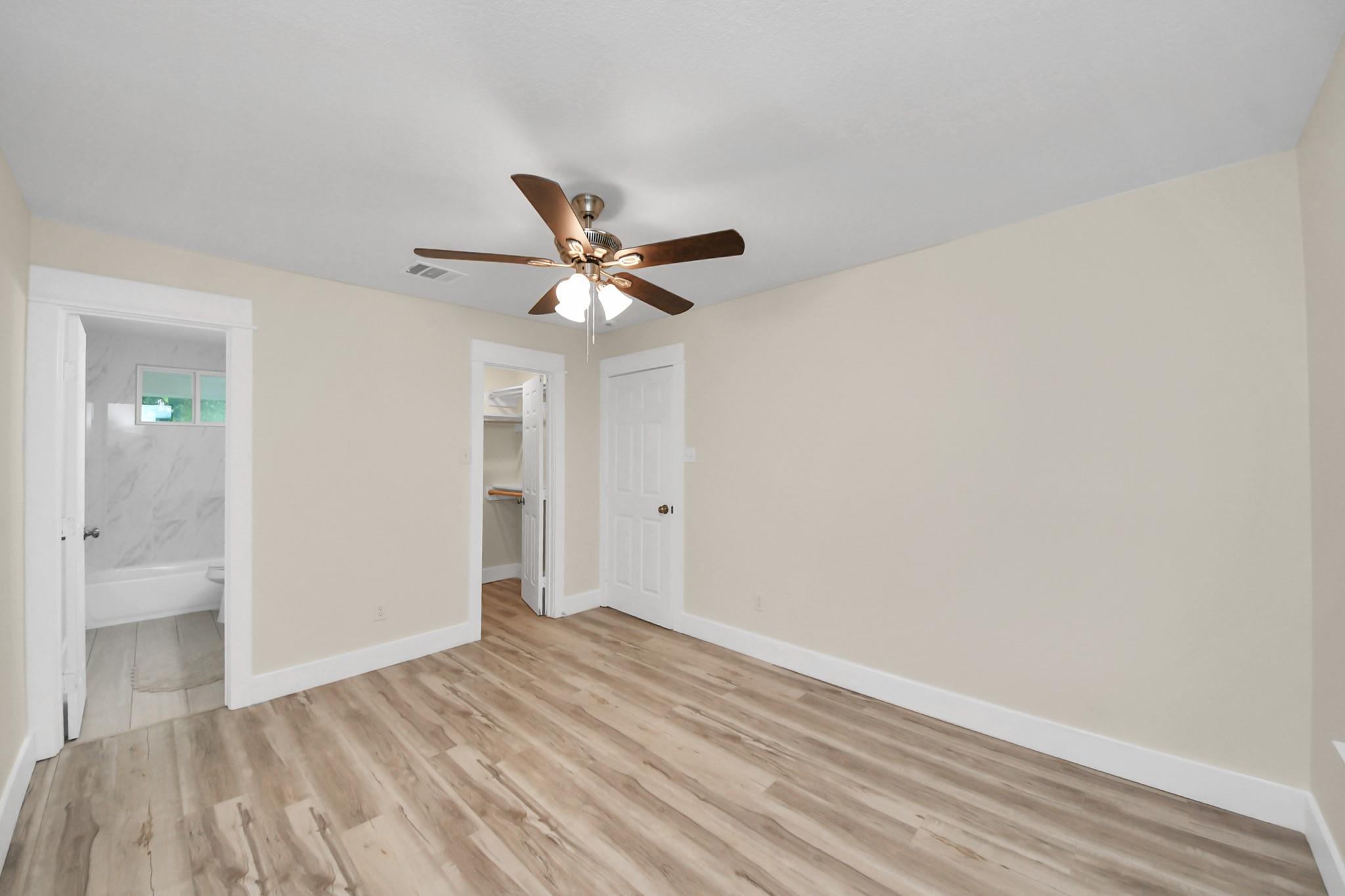 Empty bedroom with wood-look flooring and a ceiling fan, adjoining a bathroom and closet.