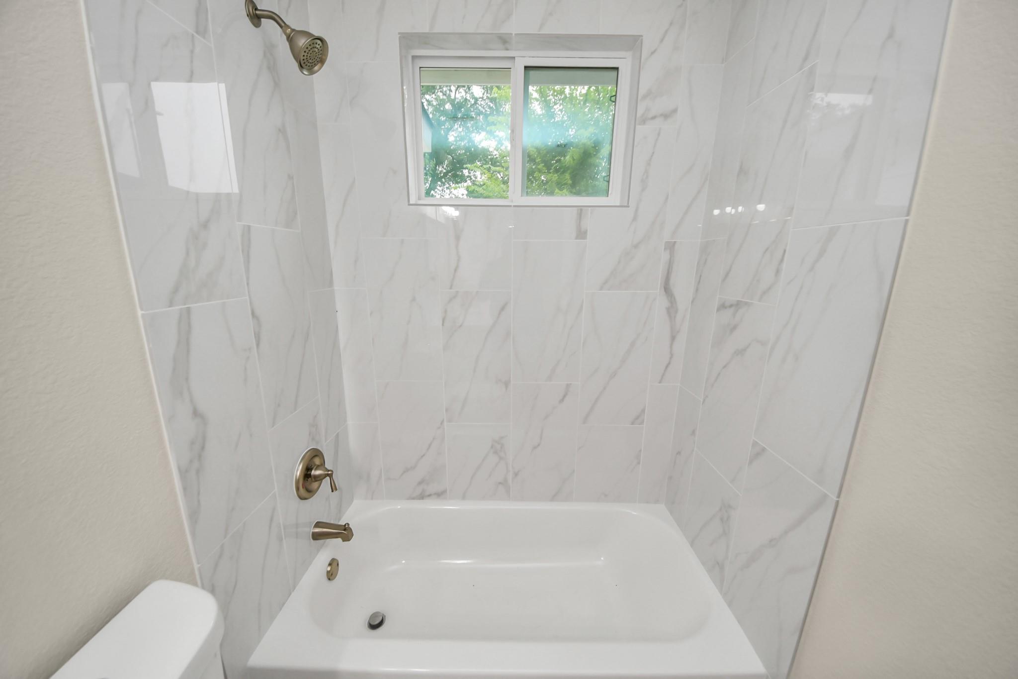 Bathroom bathtub with marble-look tile walls, brass fixtures, and a window above.